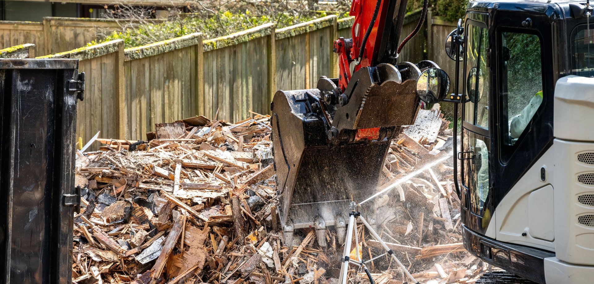 Excavator demolishing debris in a fenced yard, with dust flying from the bucket