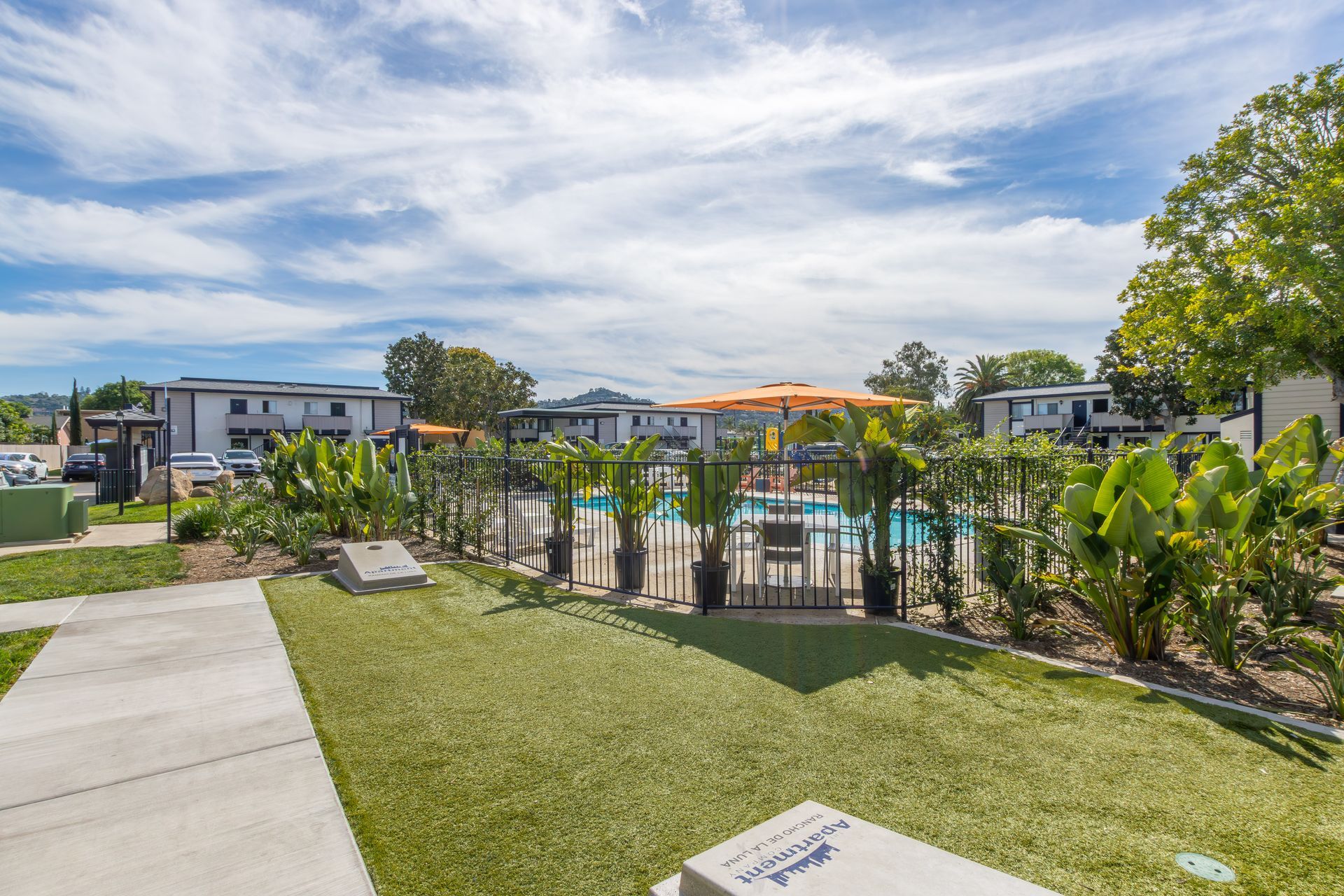Apartment complex pool with fenced deck, green lawn, and cloudy blue sky