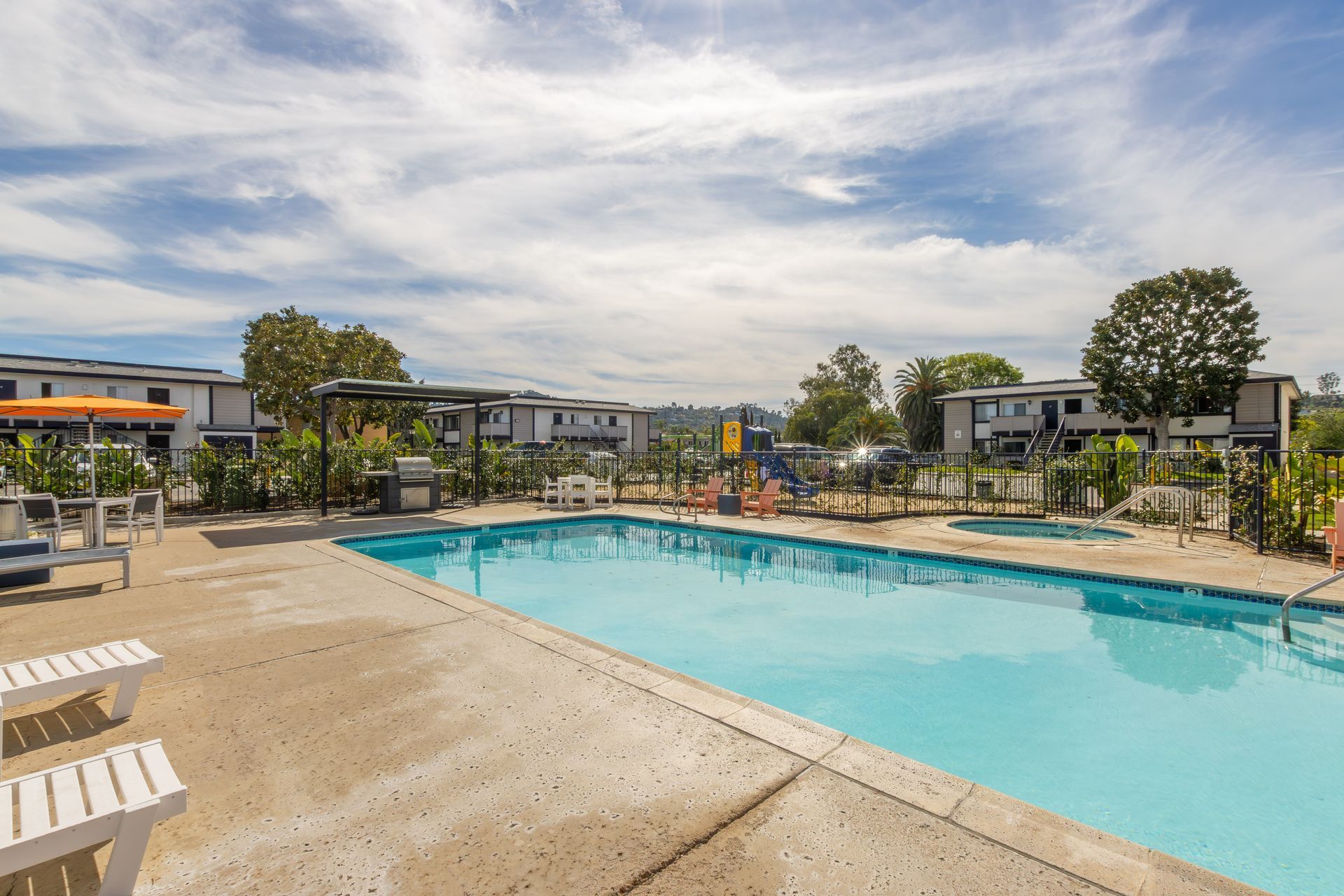Outdoor swimming pool with lounge chairs and apartment buildings under a partly cloudy sky