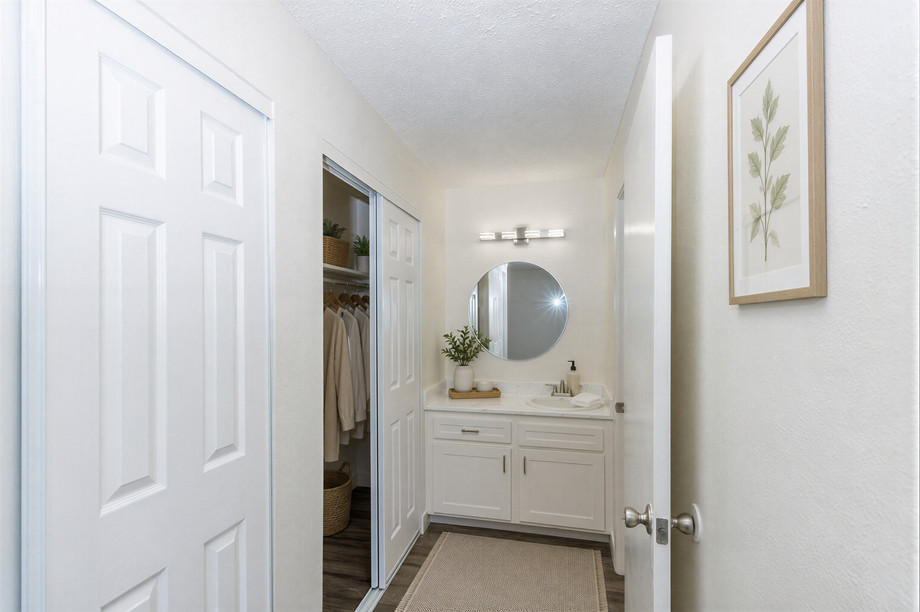 Bright bathroom hallway with white vanity, round mirror, and closet door open on the left