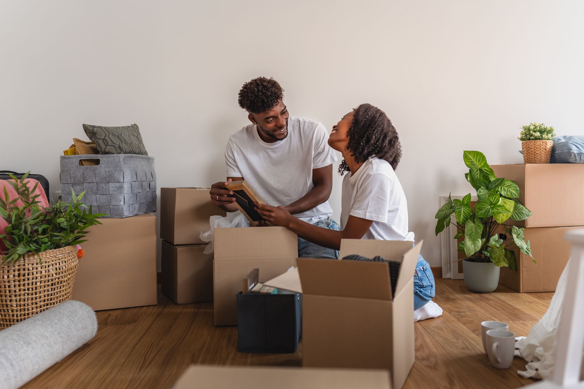 Two people sit on a wooden floor surrounded by cardboard moving boxes, looking at each other and laughing while unpacking.