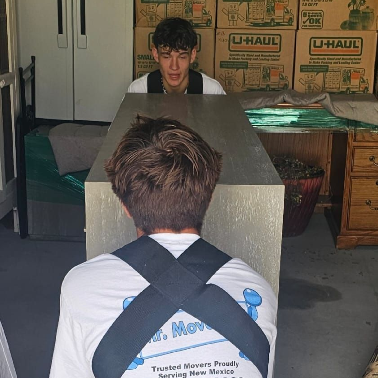 Two people wearing moving harnesses face each other while lifting a large, light-colored desk inside a room with boxes.