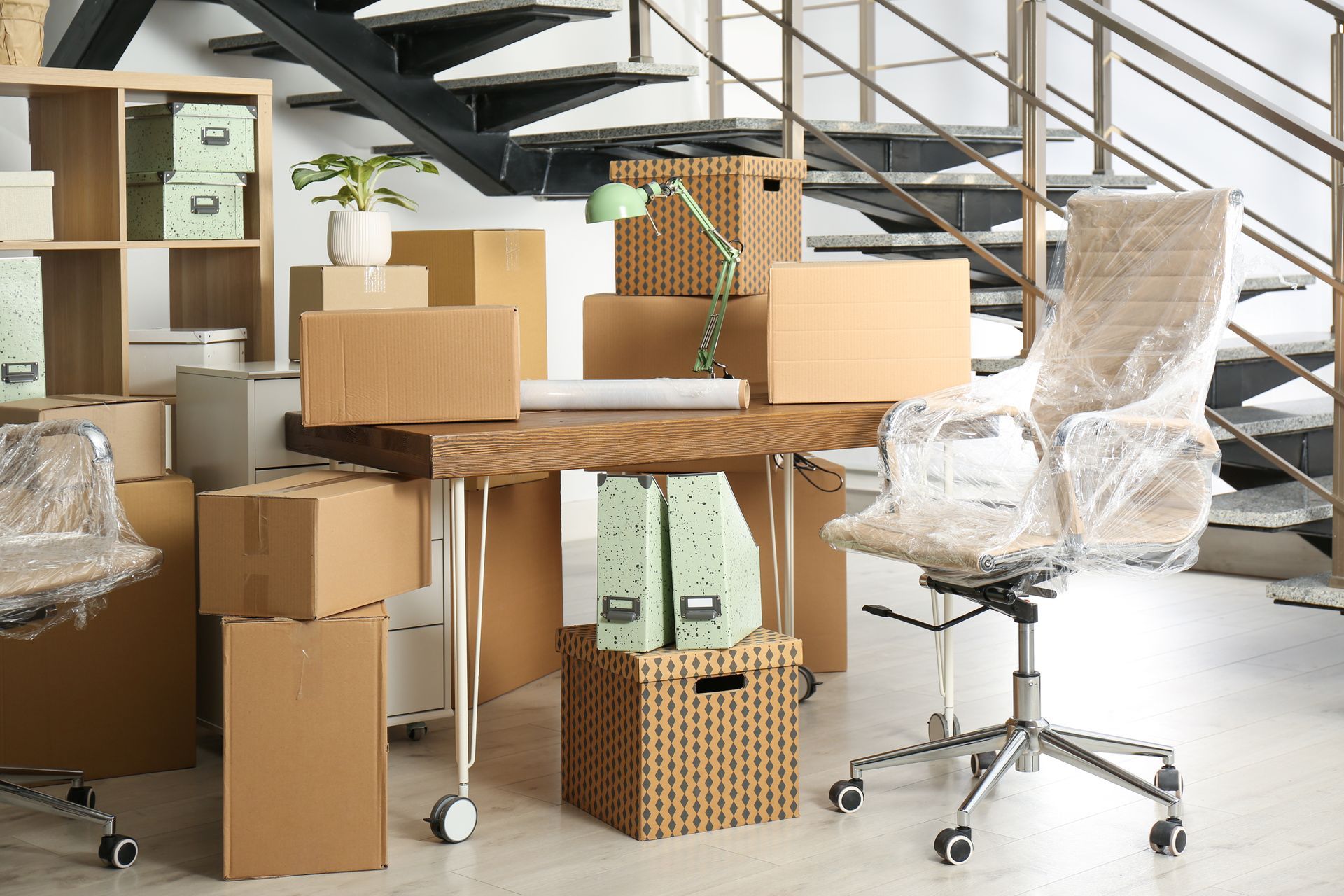 A wood desk and chair wrapped in plastic, surrounded by cardboard moving boxes and office supplies in a modern office.