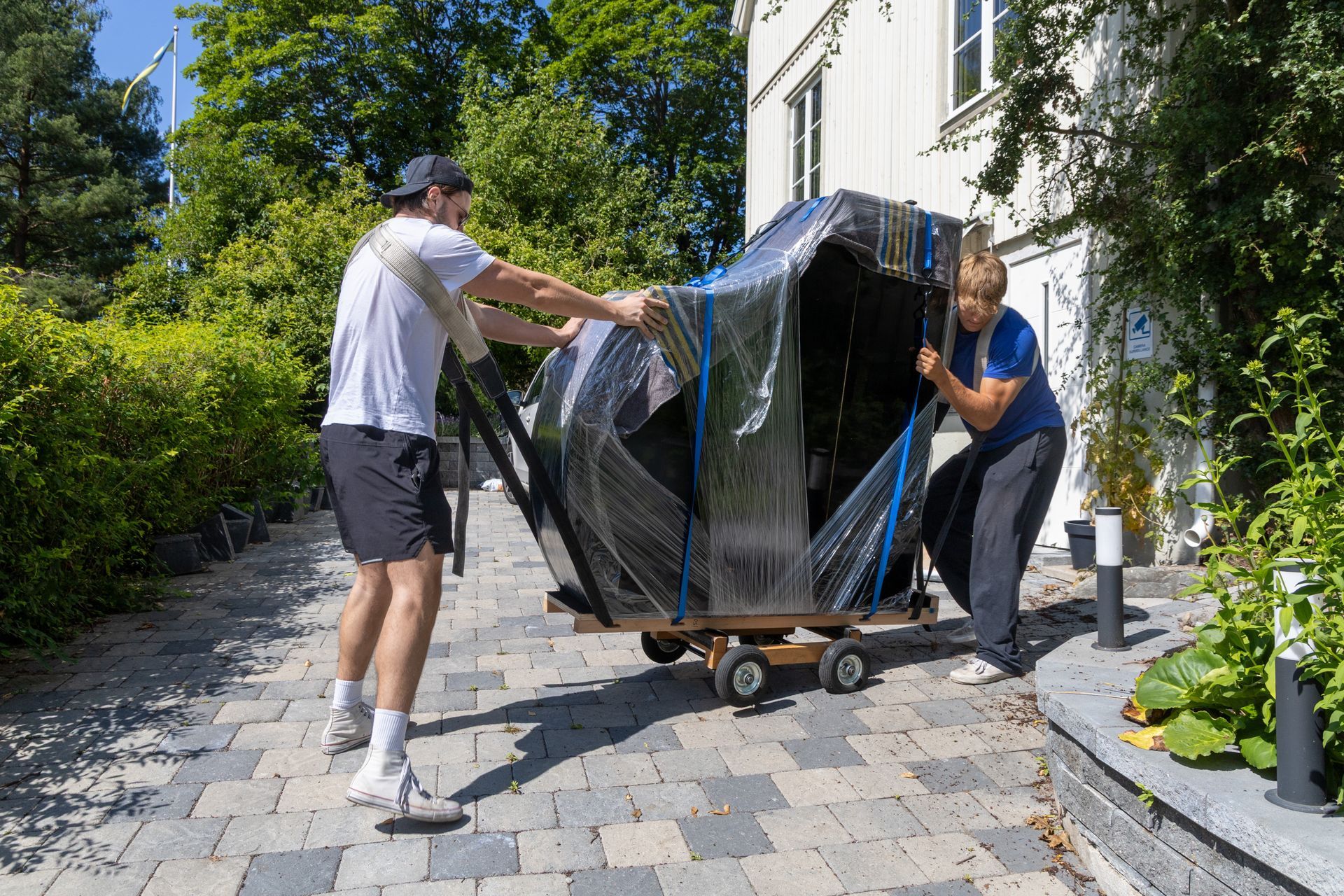 Two people pushing a wrapped piano on a dolly along a sunny stone driveway