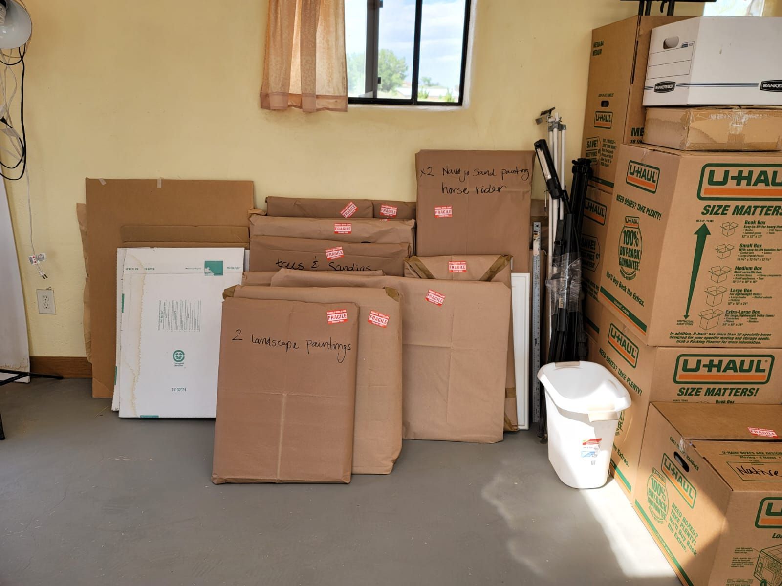 Cardboard moving boxes and flat-packed furniture items stacked against a yellow wall in a room.