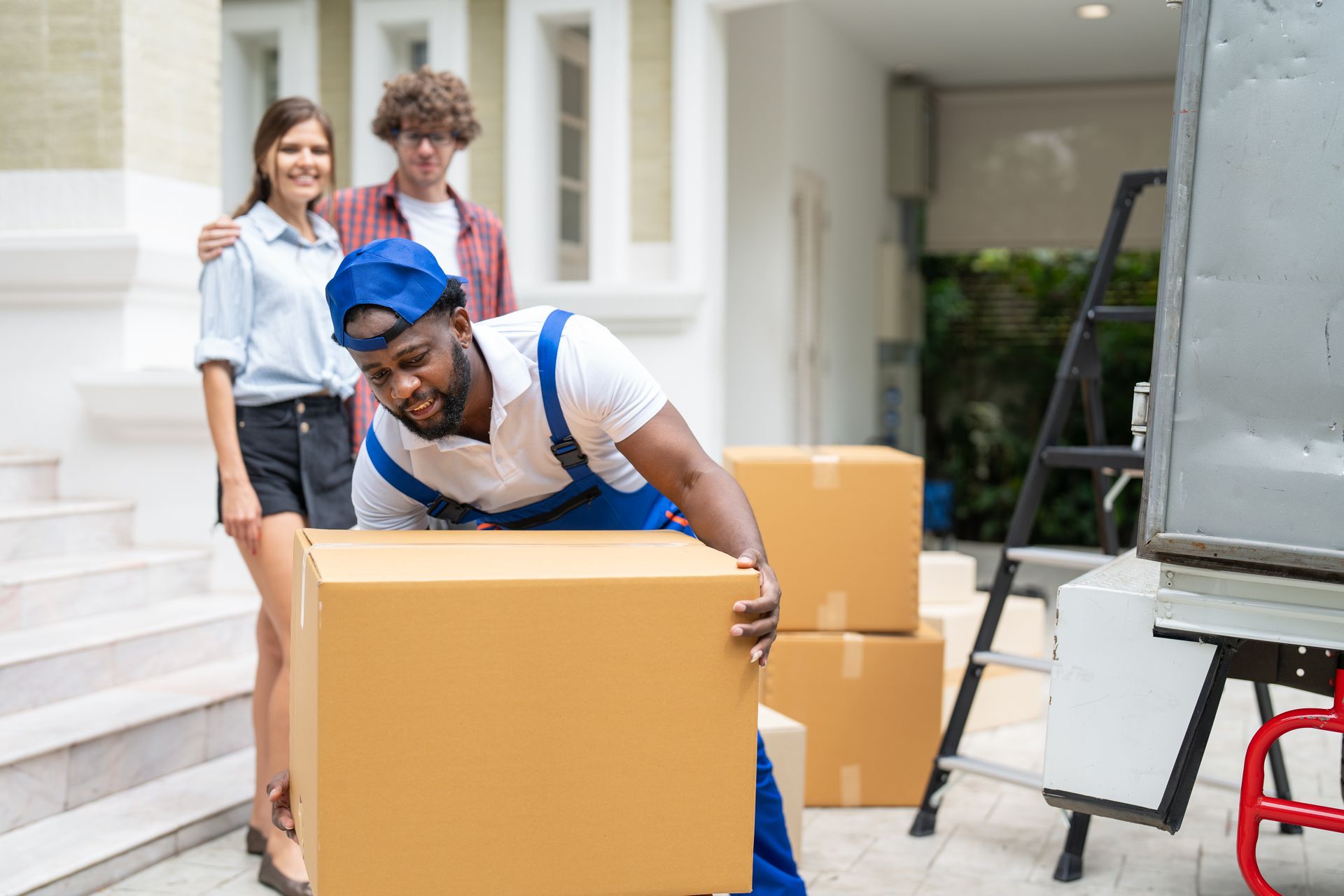 Mover carrying a large cardboard box while two people watch in a house entryway