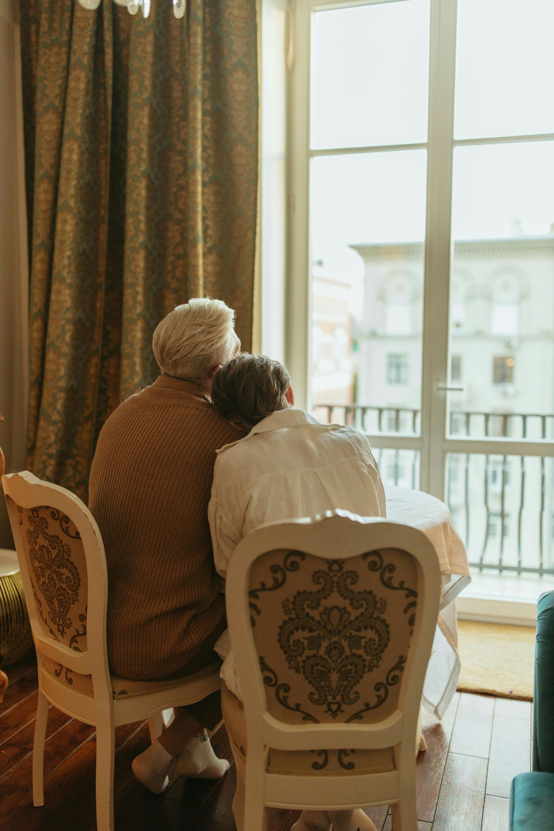 Two people seated, facing away from the camera, at a table near a window. Sunlight illuminates the room.