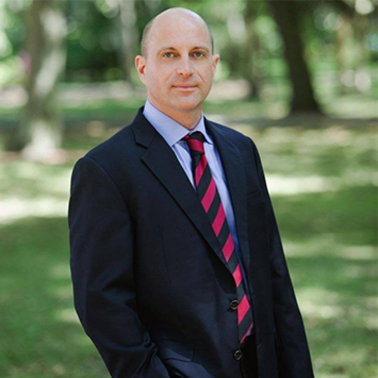 Man in a dark suit and striped tie standing outdoors in a park-like setting