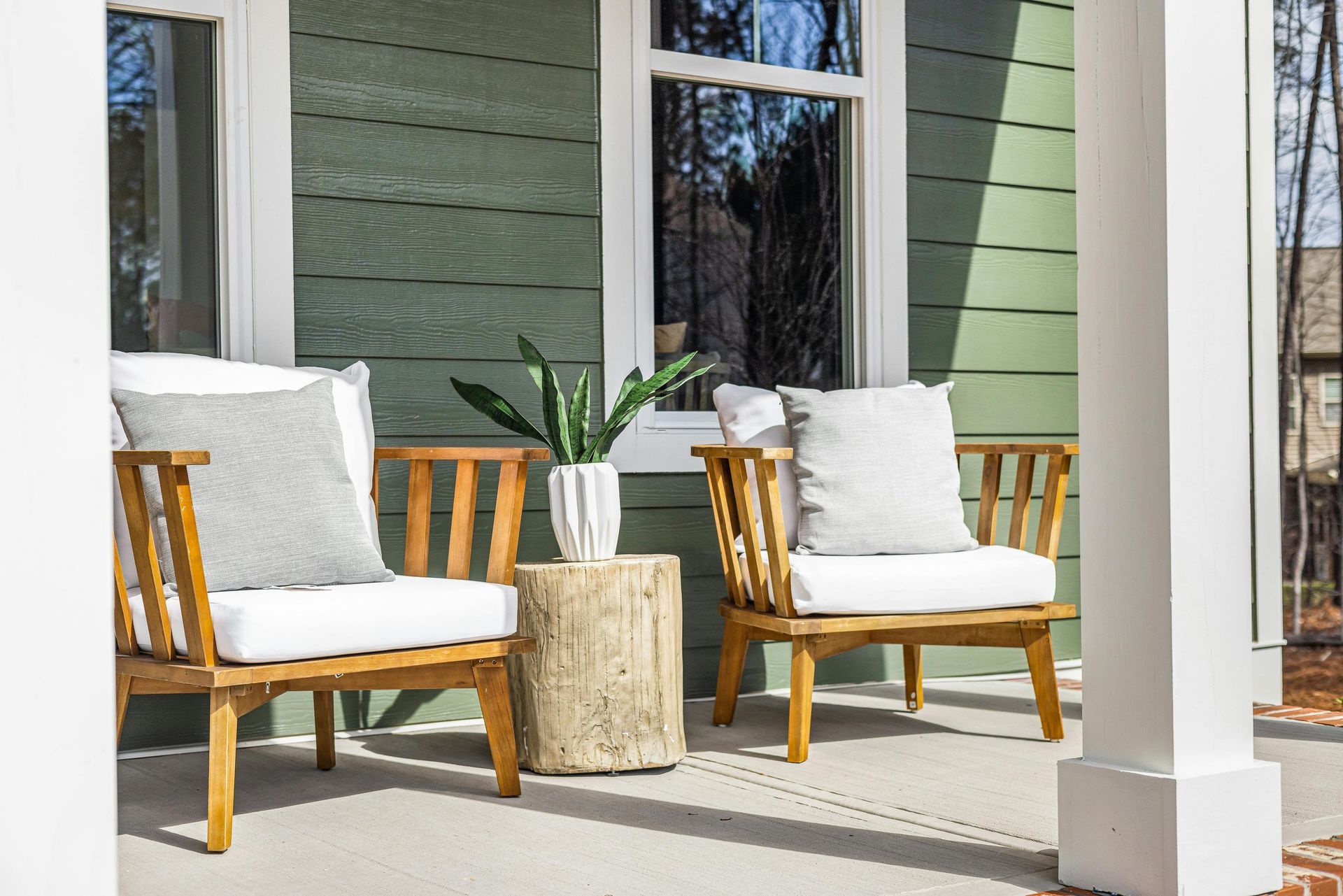 Two wooden chairs with white cushions and grey pillows sit on a porch next to a stump table with a potted plant.
