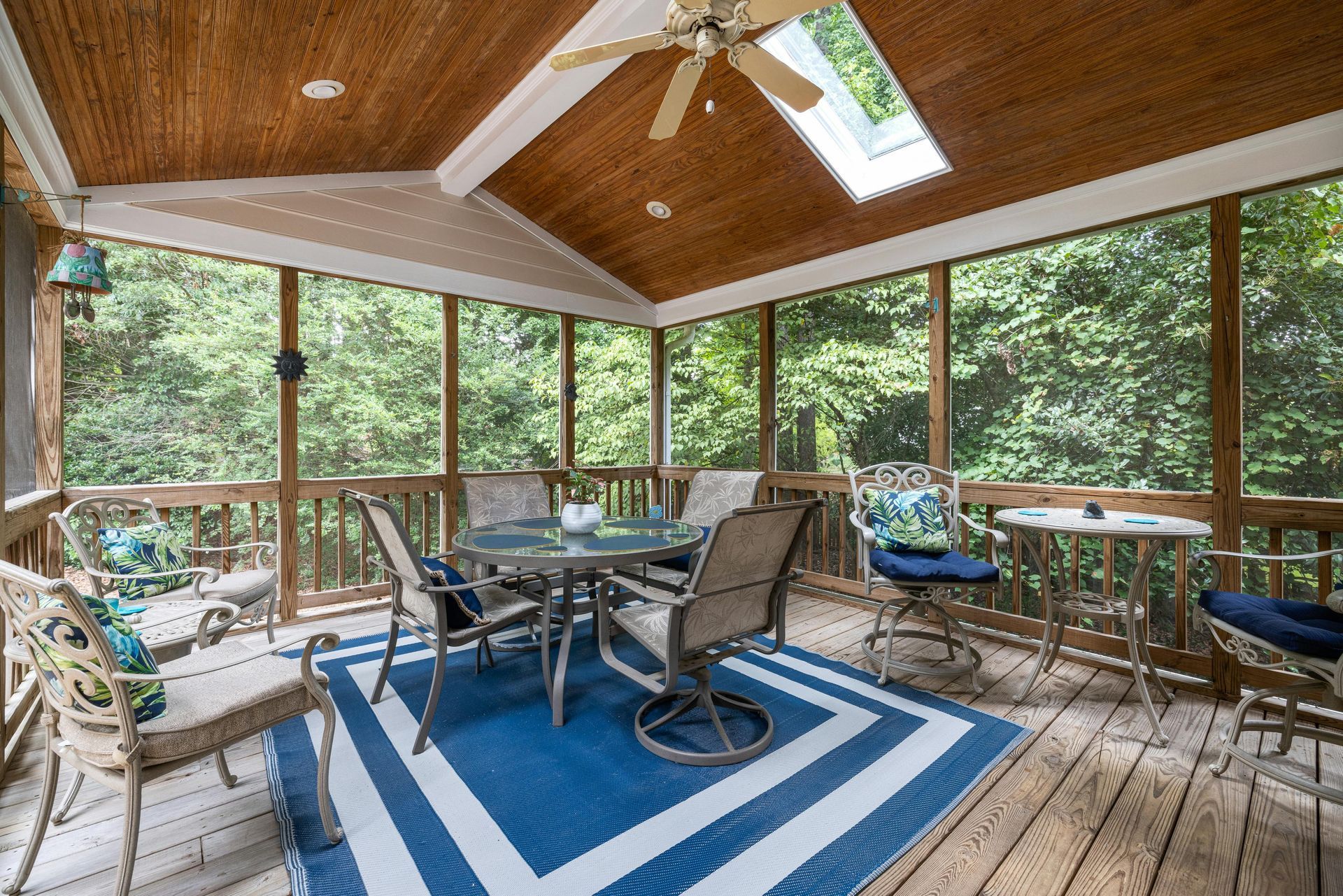 A screened-in porch with a wood-paneled ceiling, ceiling fan, skylight, and outdoor furniture on a blue striped rug.