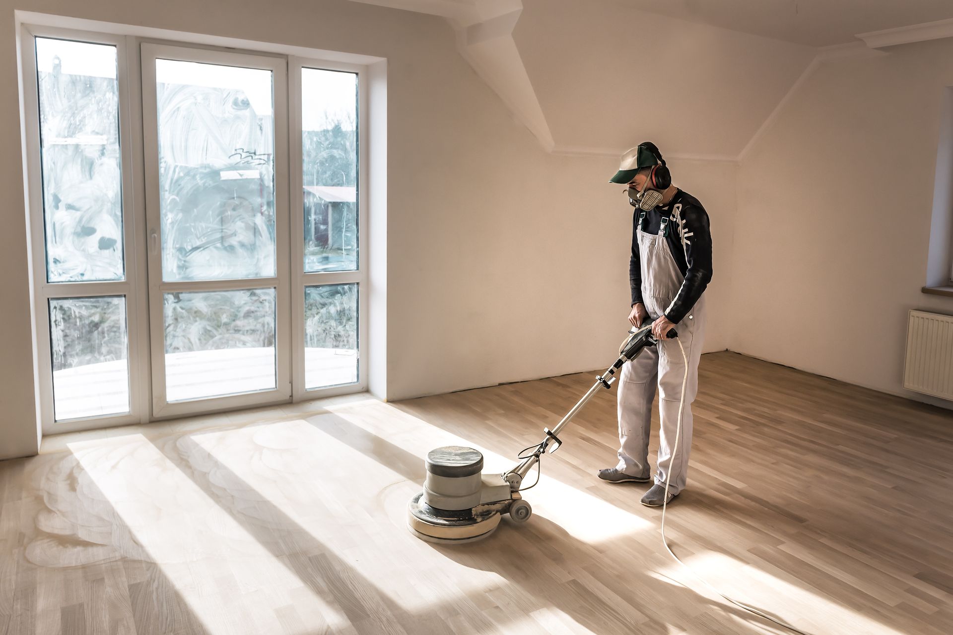 Person sanding a wooden floor in a sunlit room, wearing a respirator and overalls.