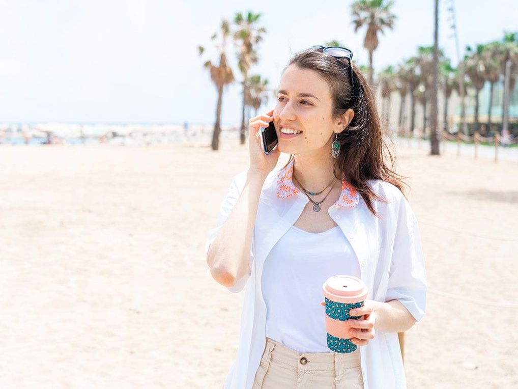 A woman is talking on a cell phone while holding a cup of coffee on the beach.
