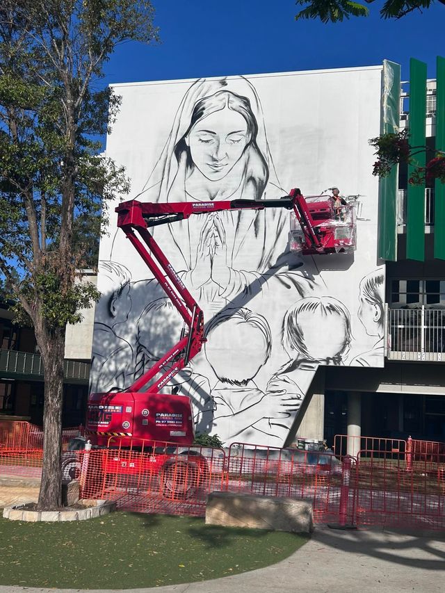 Cherry-picker painting a large black-and-white mural on a building exterior under a clear blue sky.