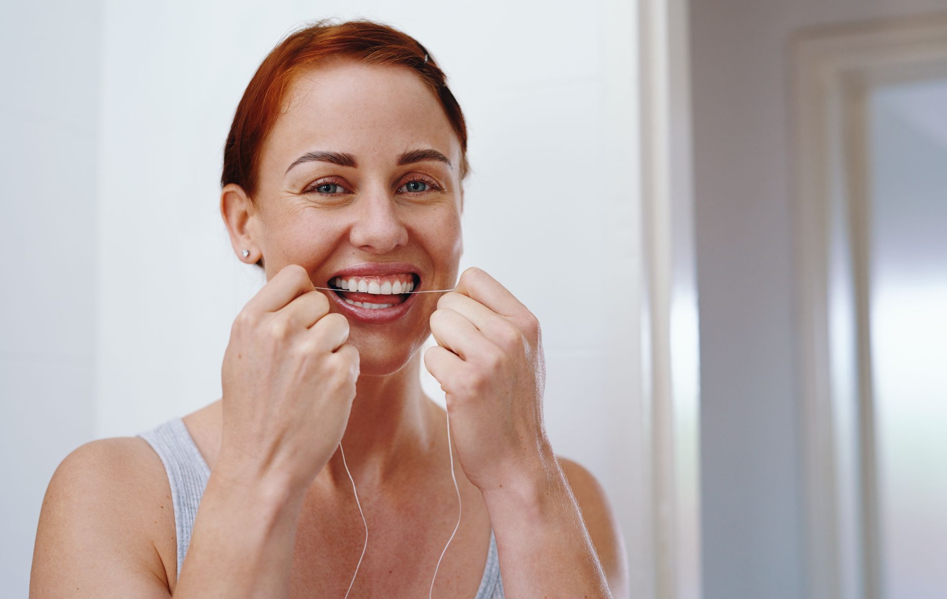 Woman flossing teeth in a bright bathroom, smiling at the mirror