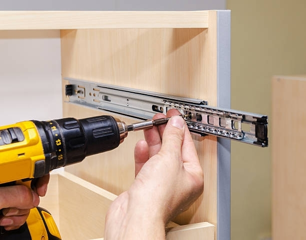 A person is using a drill to install drawer slides on a cabinet.