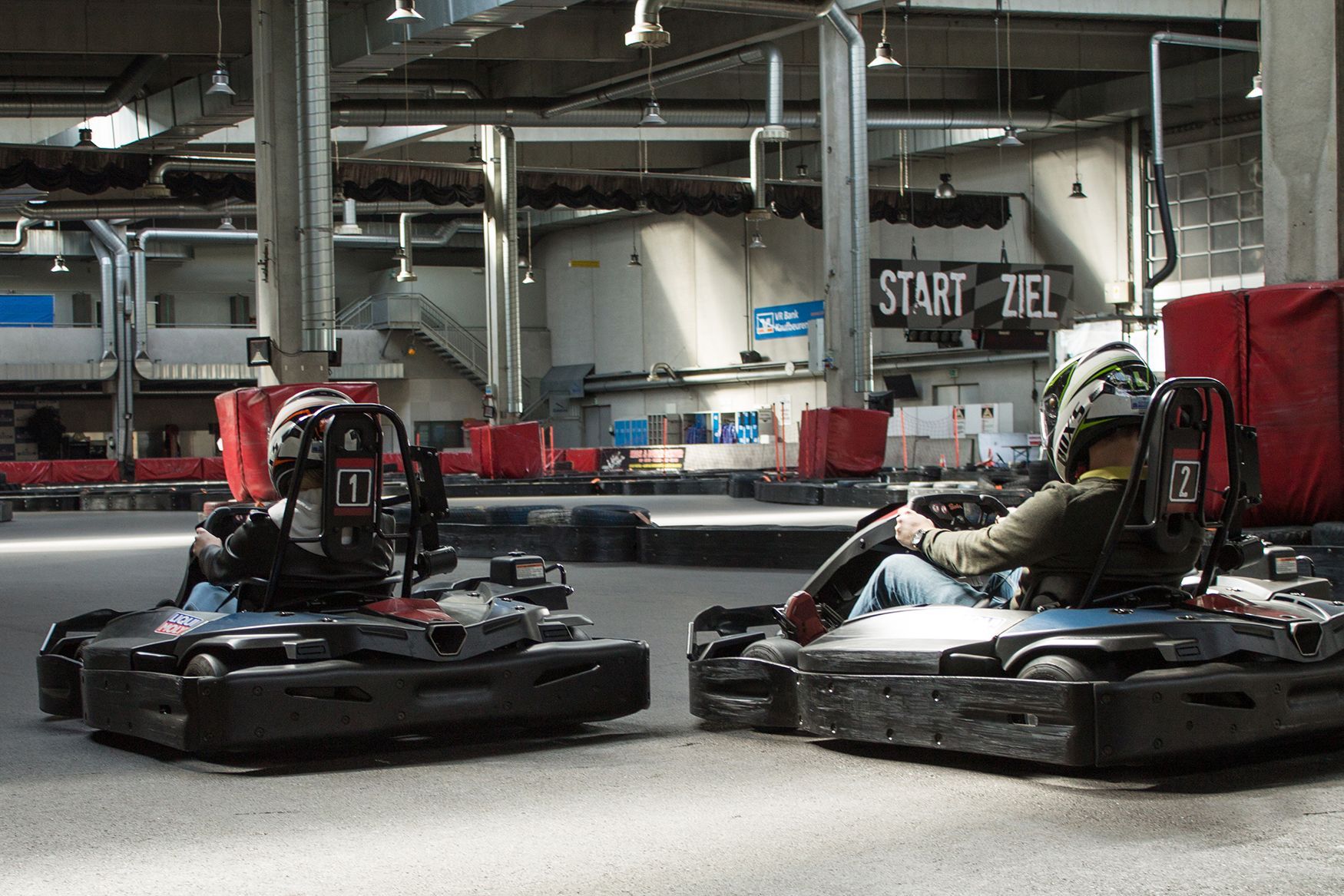 Zwei Personen fahren in schwarzen Go-Karts in einer Halle um die Wette; im Hintergrund ist ein START/ZIEL-Schild zu sehen.
