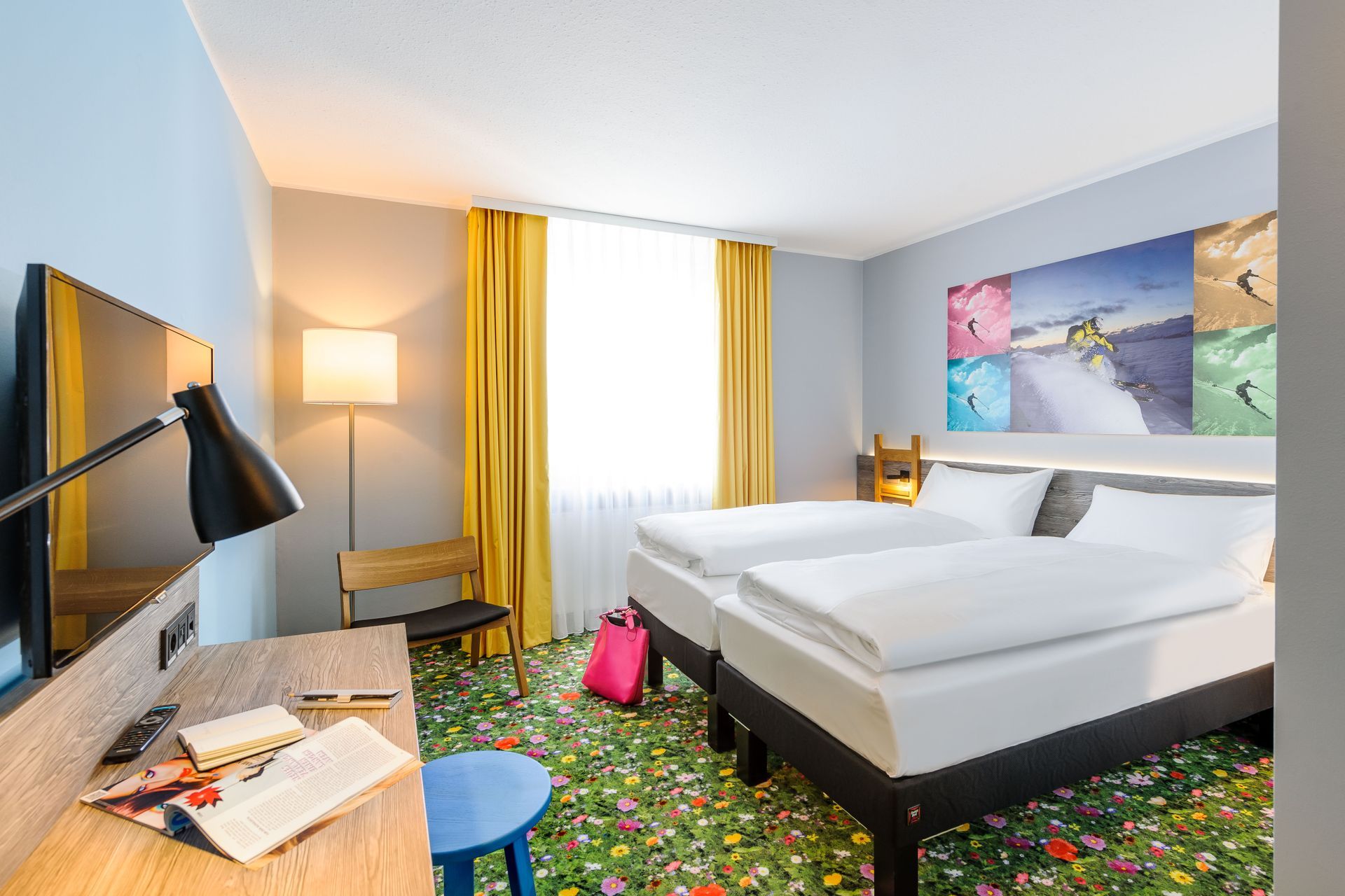 Modernes Hotelzimmer mit zwei Betten, farbenfrohem Teppich, Schreibtisch, Lampe und Wandbild