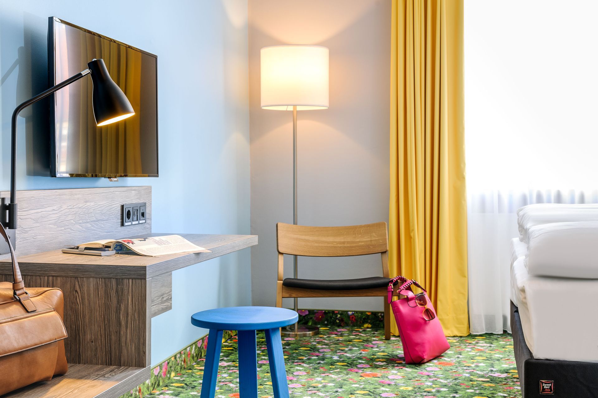 Modernes Hotelzimmer mit Bett, Schreibtisch, Stehlampe, gelben Vorhängen und blauem Hocker