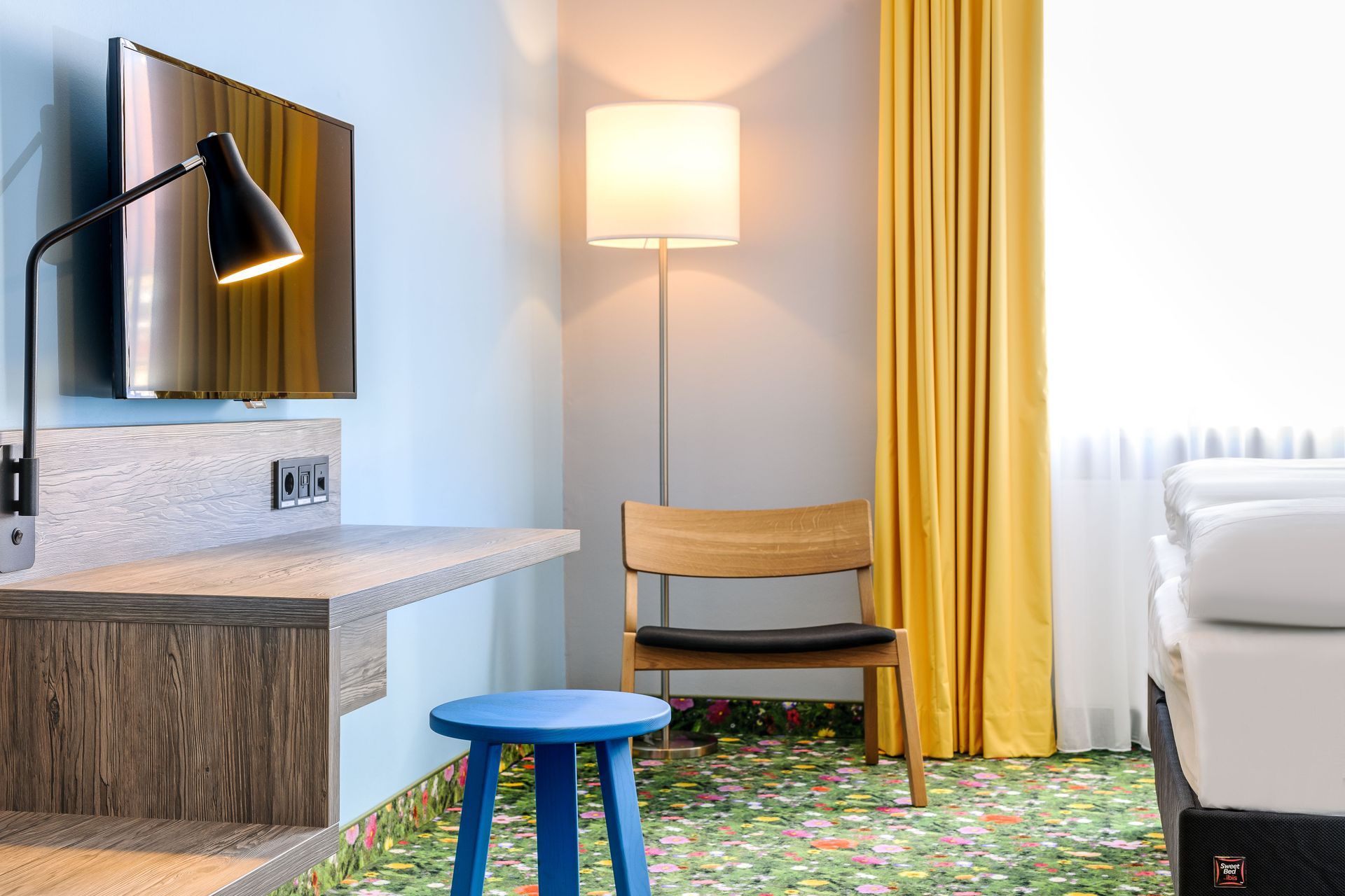 Modernes Hotelzimmer mit Bett, Stehlampe, blauem Hocker, Schreibtisch, Spiegel und gelben Vorhängen
