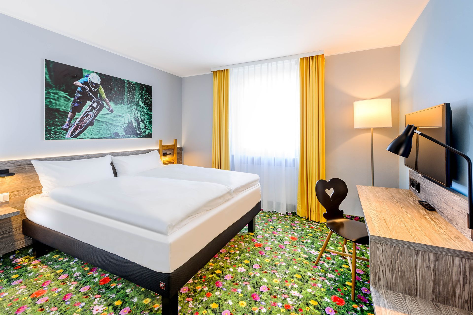Modernes Hotelzimmer mit Bett, geblümtem Teppich, Wandbildern, Schreibtisch, Lampe und großem Fenster mit Vorhängen.