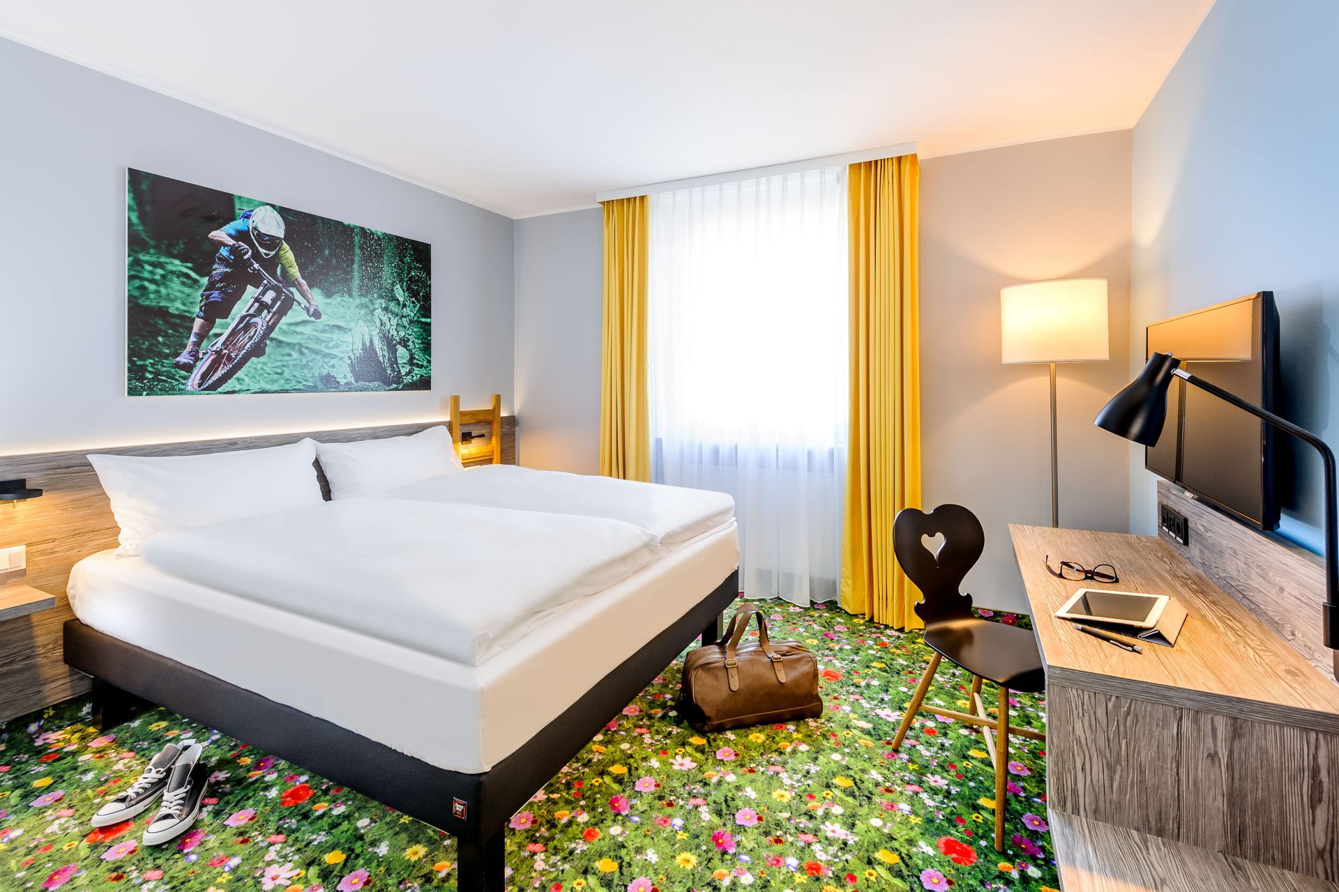 Helles Hotelzimmer mit Bett, Schreibtisch, Fernseher und grünem Teppich mit Blumenmuster.