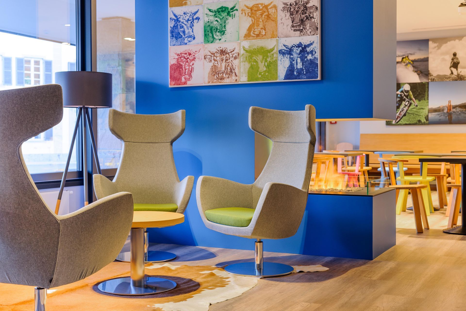 Moderne Bürolounge mit grauen Stühlen, blauen Wänden, farbenfrohen Kunstwerken und einem hellen Holztischbereich