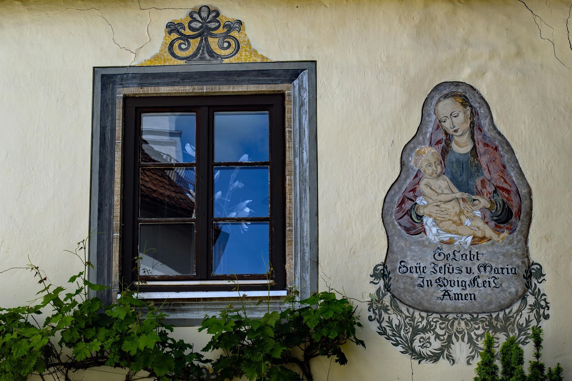 Eine Wand mit einem gerahmten Fenster und einem religiösen Wandgemälde, darunter Efeu und im Glas gespiegelter blauer Himmel.