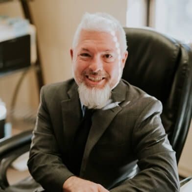 A man in a suit and tie is sitting in an office chair and smiling.