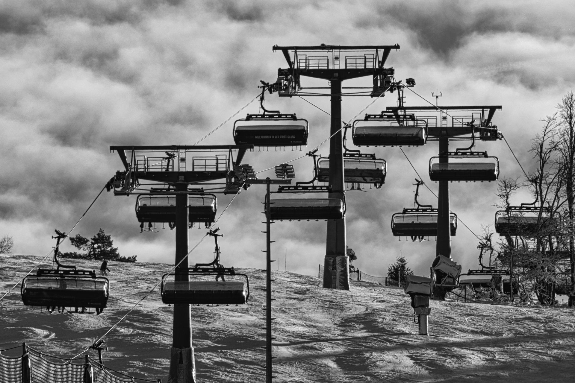 Black-and-white ski lift chairs hanging over a snowy mountain slope under cloudy skies