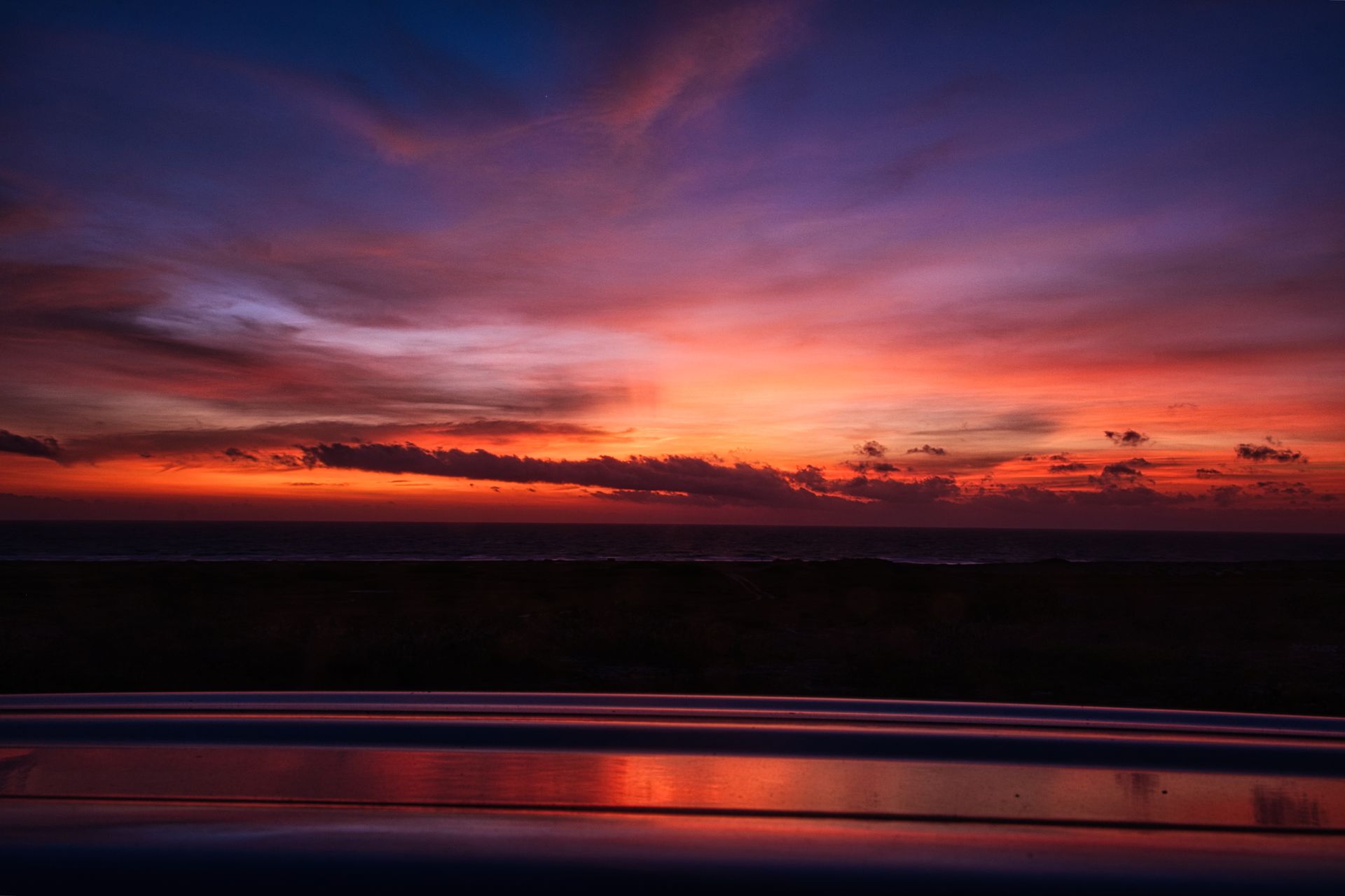 Vibrant purple and orange sunrise over calm water with reflections and a dark shoreline