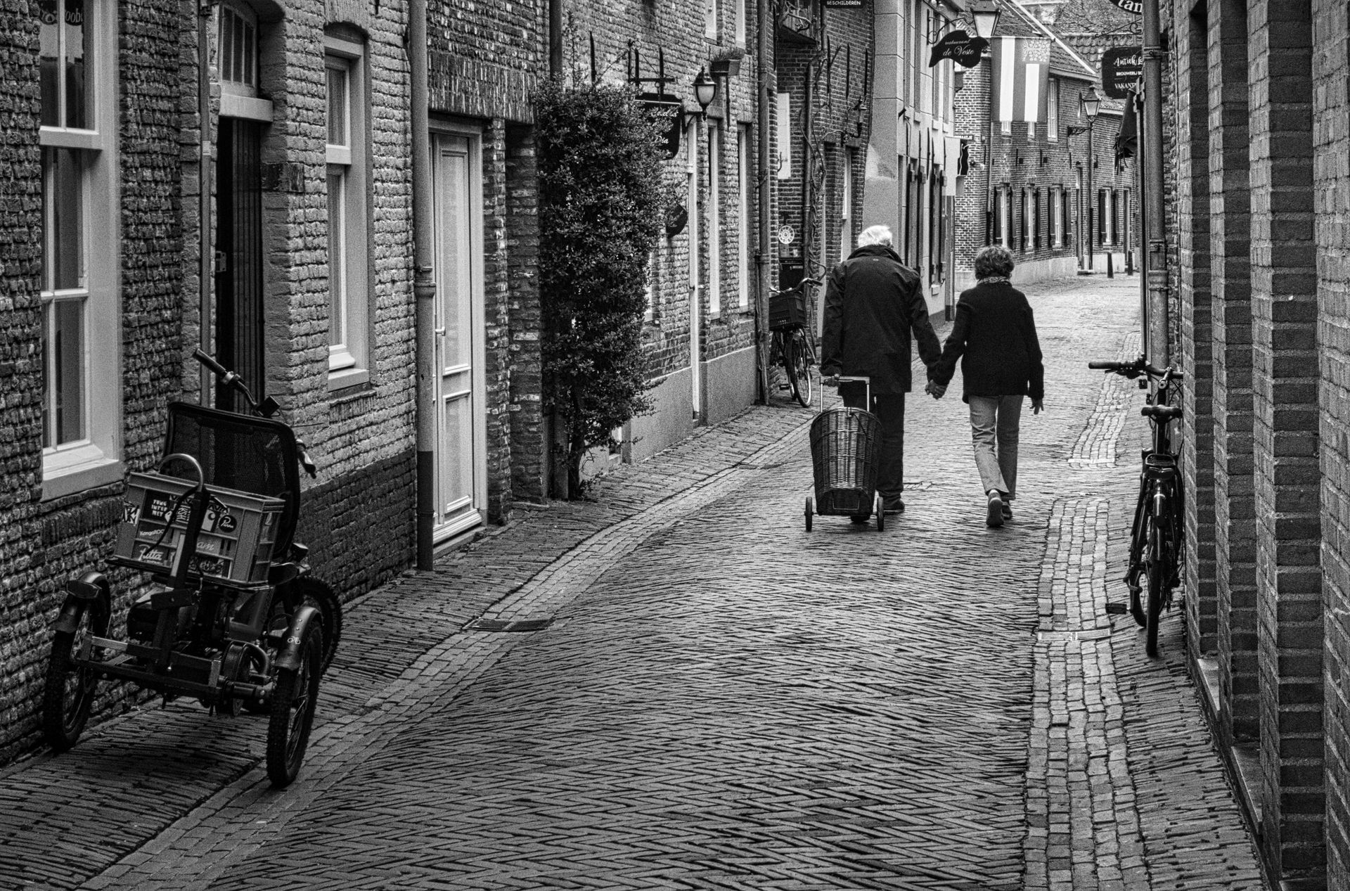 Two people walking down a narrow cobblestone alley, one pulling a wheeled bag, with brick buildings on both sides