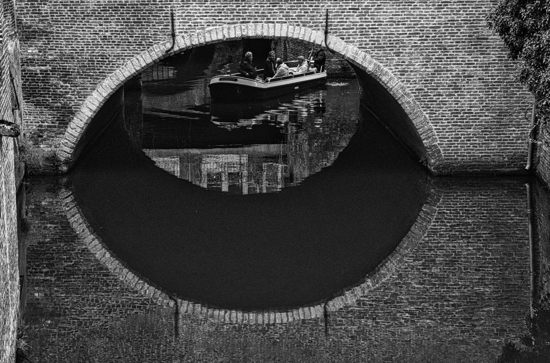 Black-and-white view of a brick arch bridge over water, with a boat reflected beneath the arch