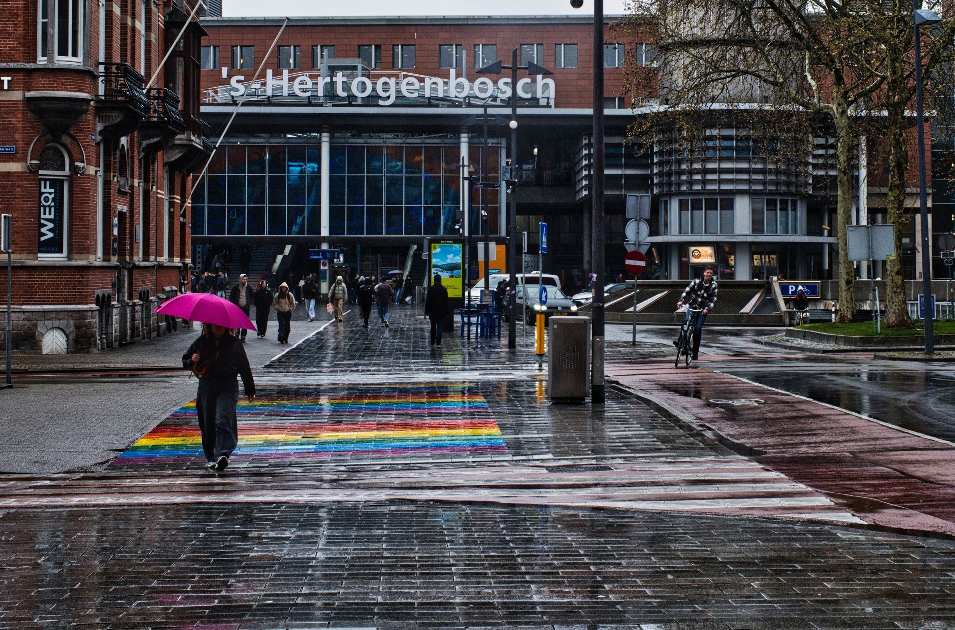 Rainy city square outside Station 's-Hertogenbosch, with pedestrians and a rainbow-painted crosswalk