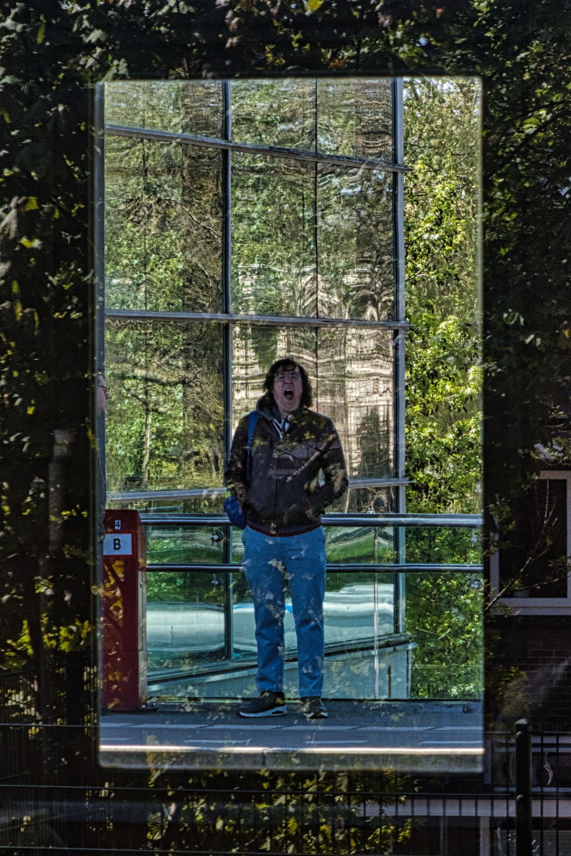 Person in camouflage jacket and jeans standing inside a glass enclosure amid a wooded outdoor setting.
