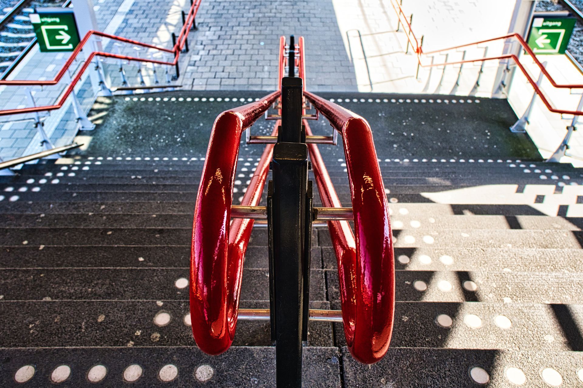 Red staircase handrails leading down a bright metal stairwell with green exit signs on both sides