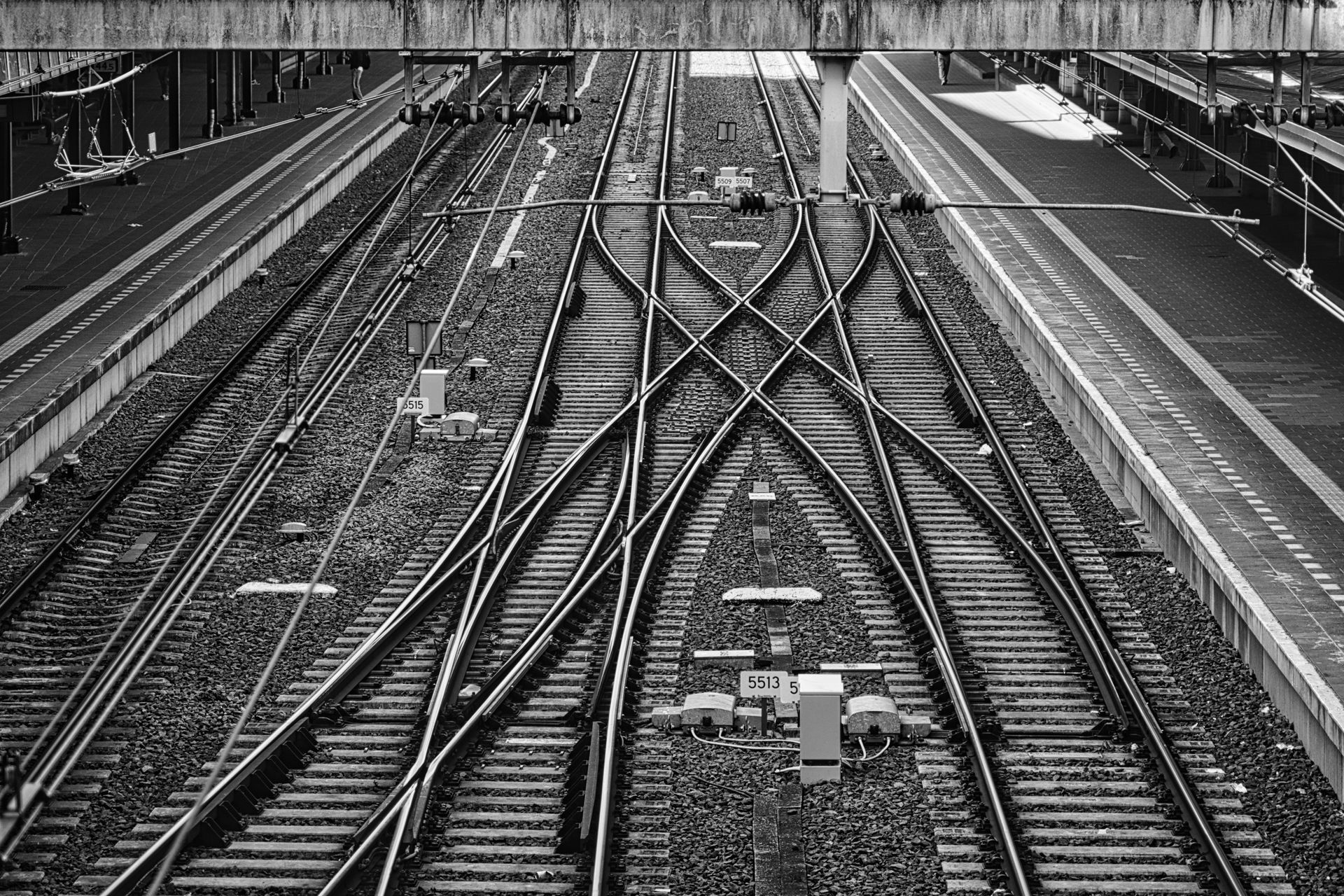 Black-and-white view of railway tracks crossing and merging beneath an overpass.