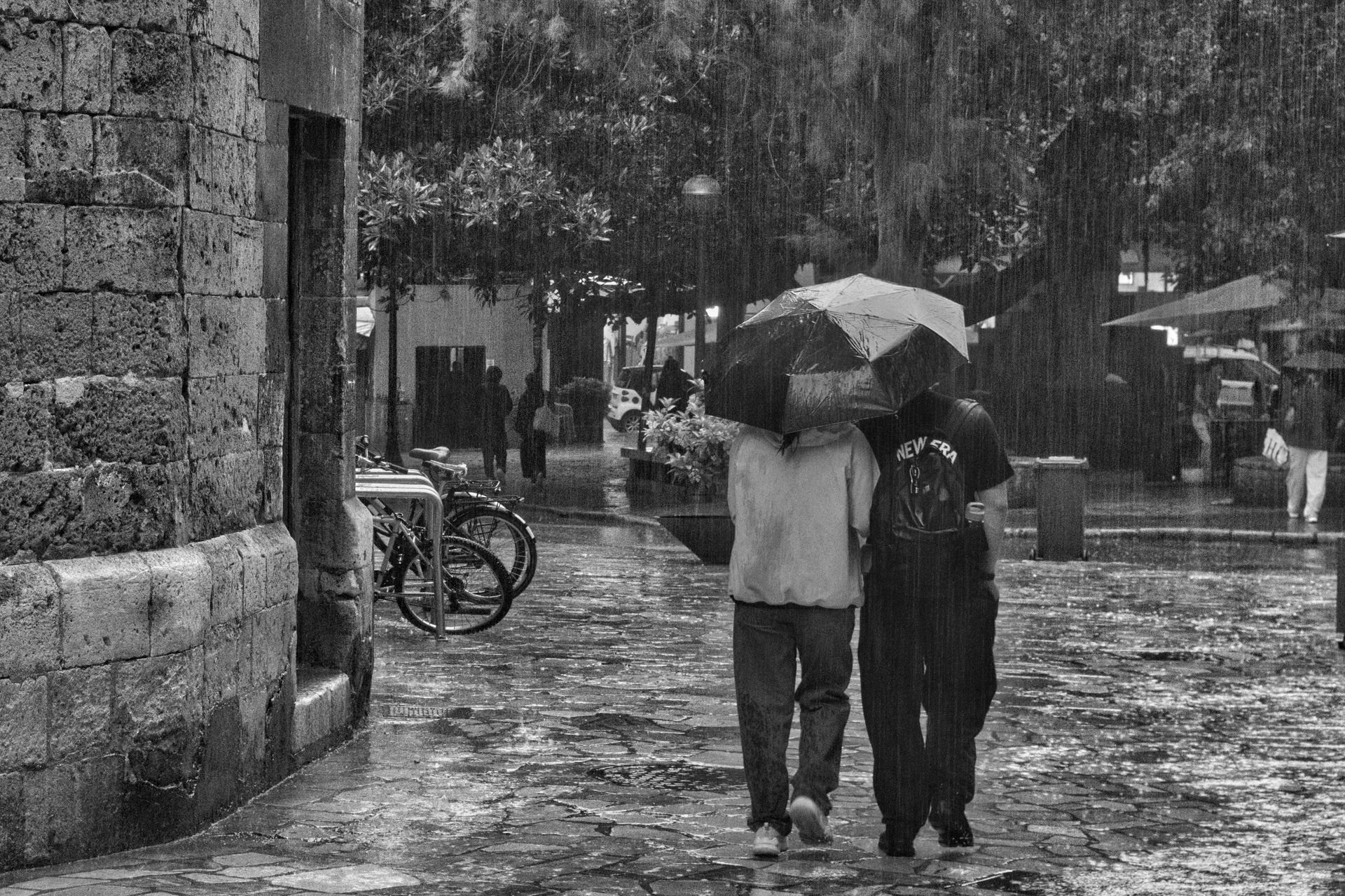 Two people walk under an umbrella in heavy rain on a cobblestone street near stone buildings.