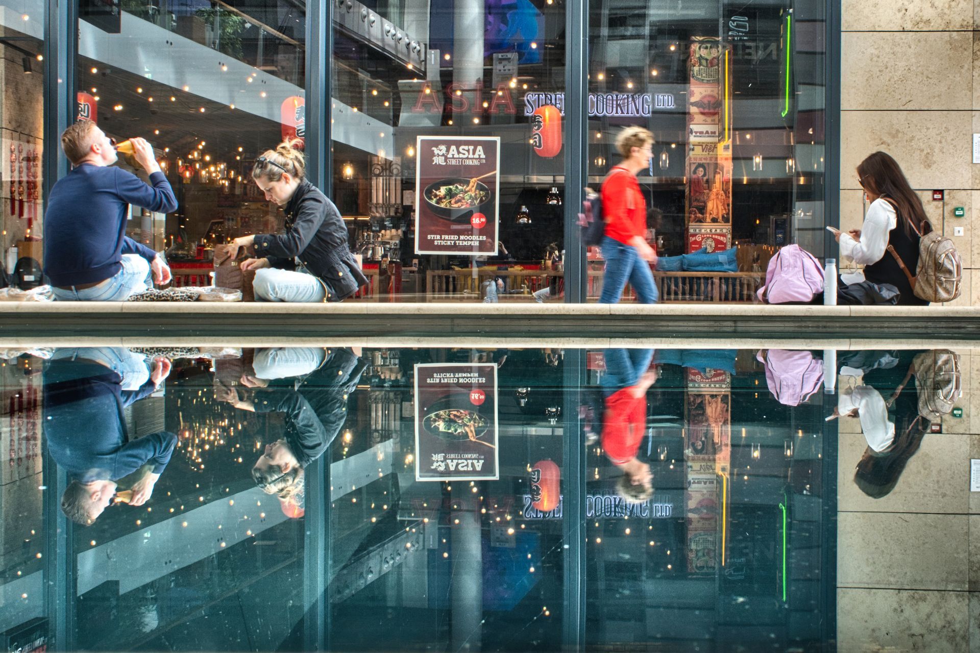 People sitting outside a café beside a reflective pool, with string lights and city reflections.
