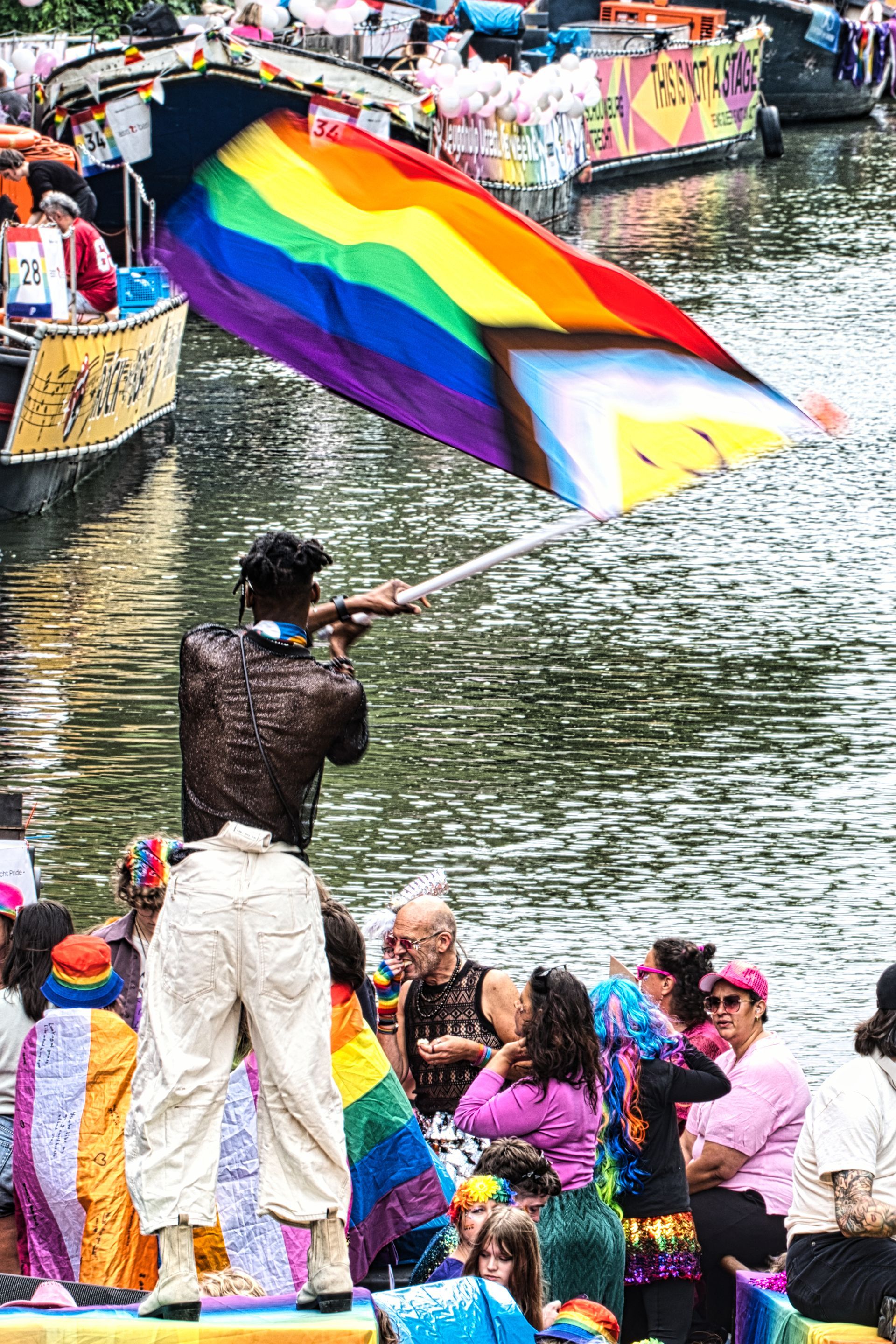Person waving a rainbow flag on a boat at a colorful waterfront celebration