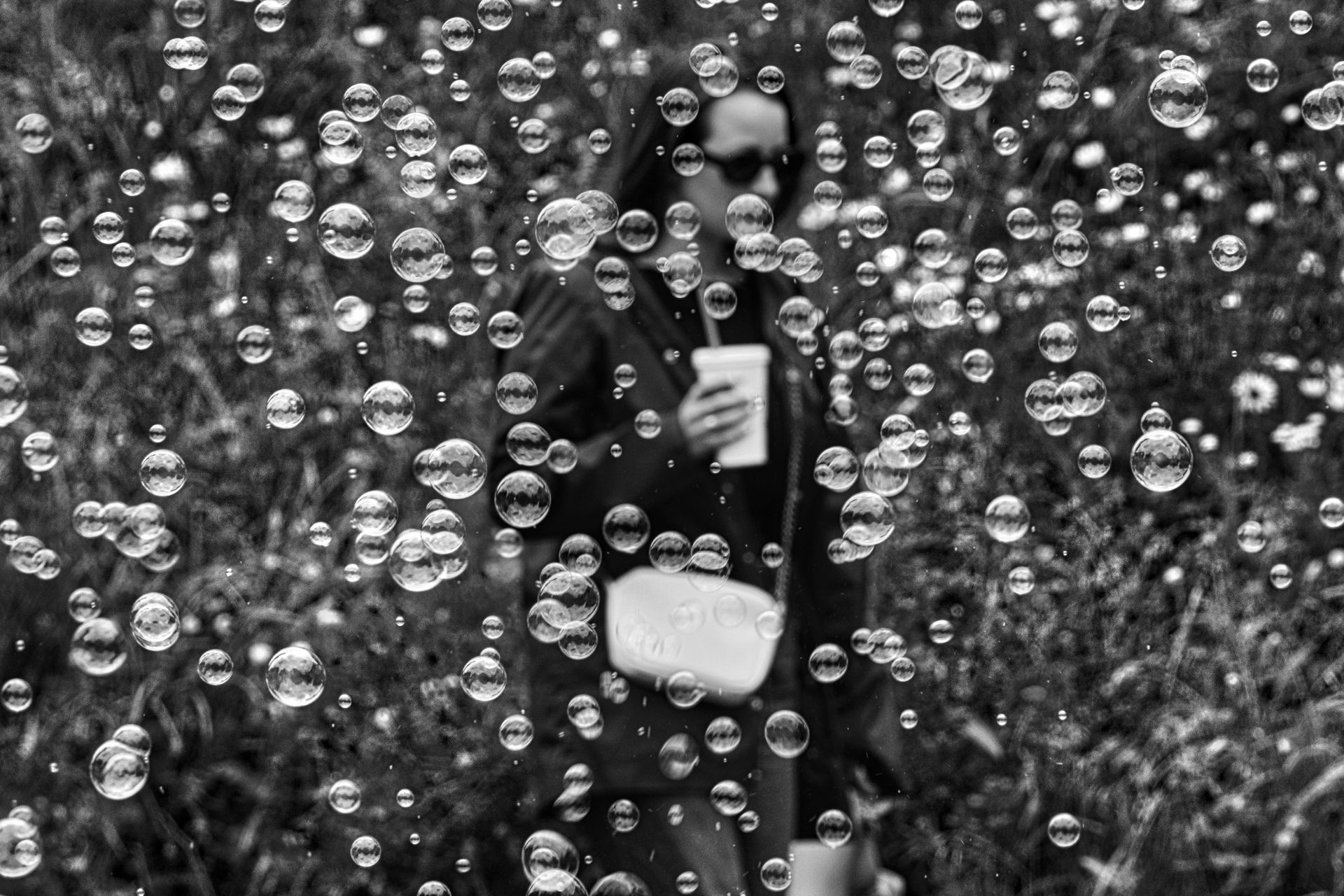 Person in black coat holding a cup, seen through a curtain of hanging beads outdoors.