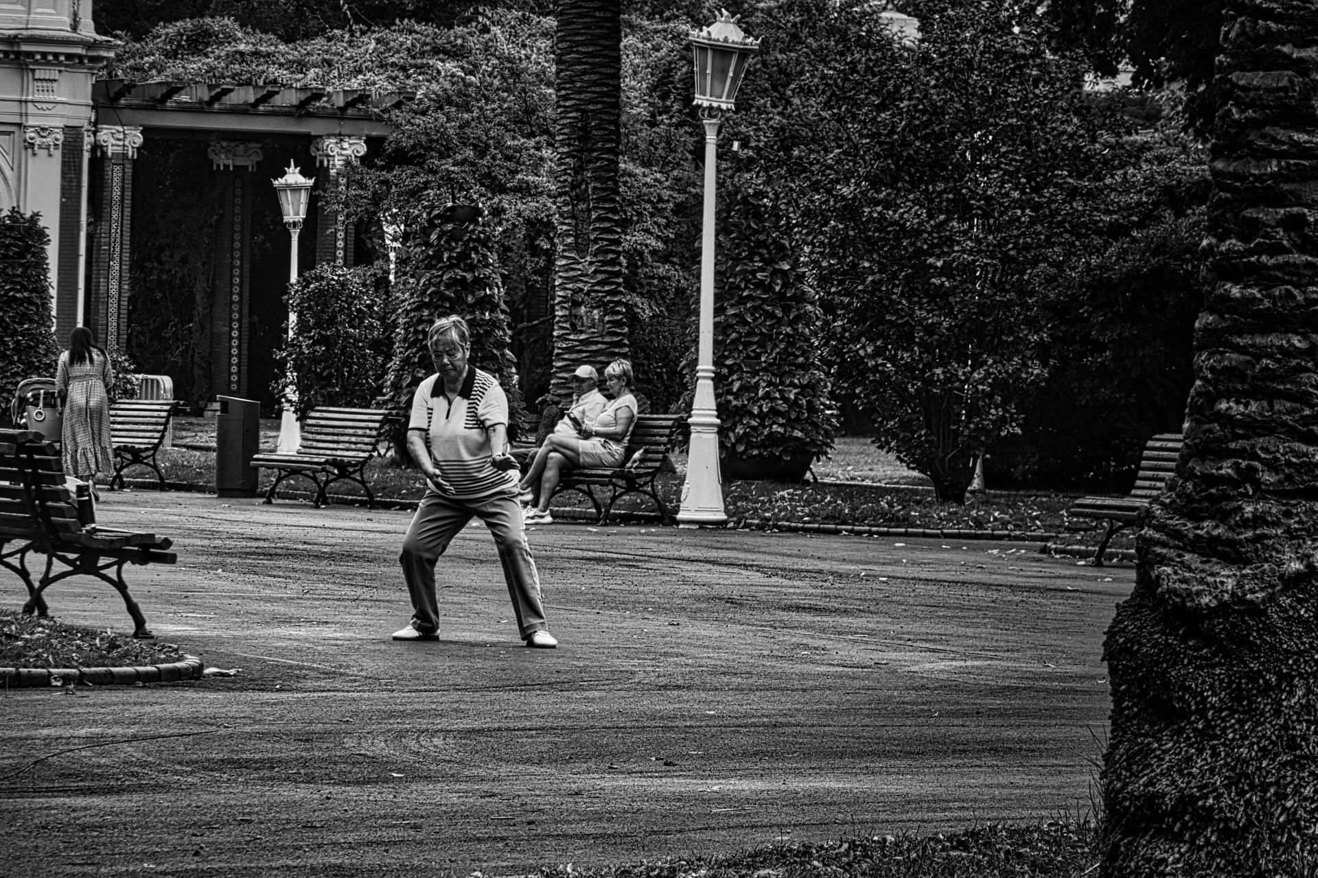 Two people standing in a park near benches and a lamppost, one holding a child