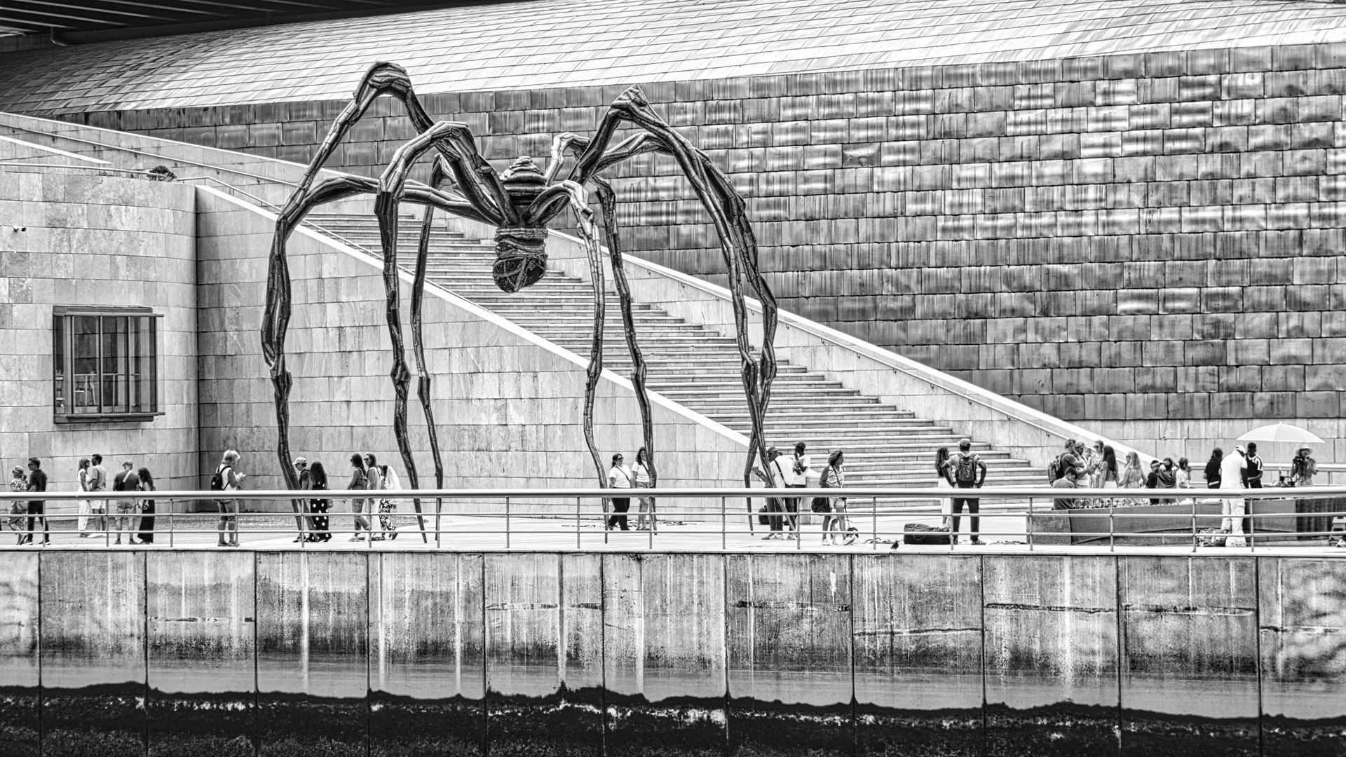 Large spider-like sculpture on a waterfront promenade with people walking beneath it, black-and-white.