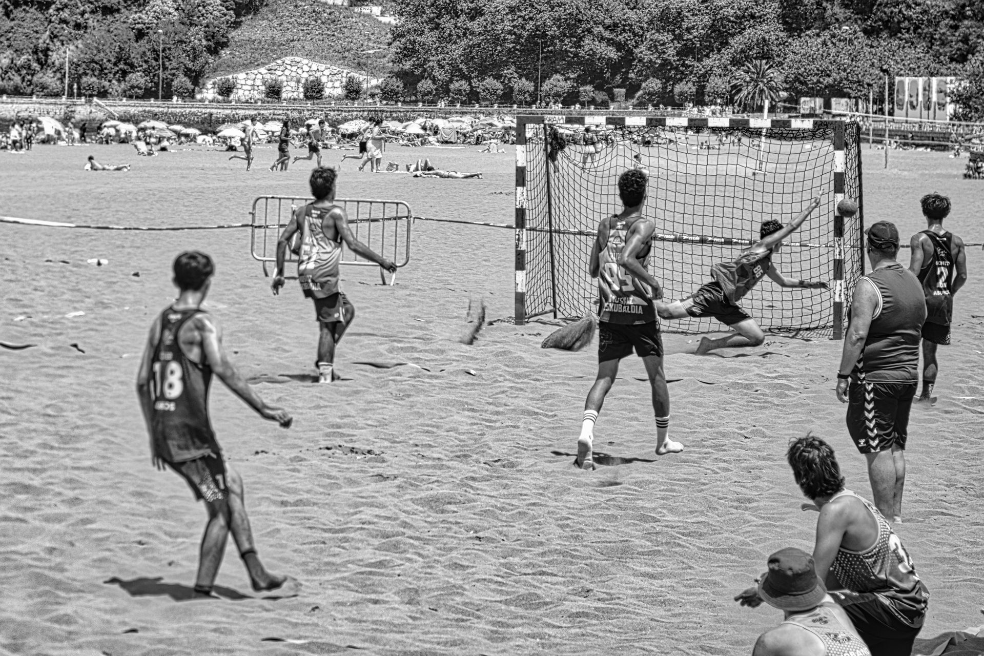 Beach handball players around a goal, with one player diving to save a shot.