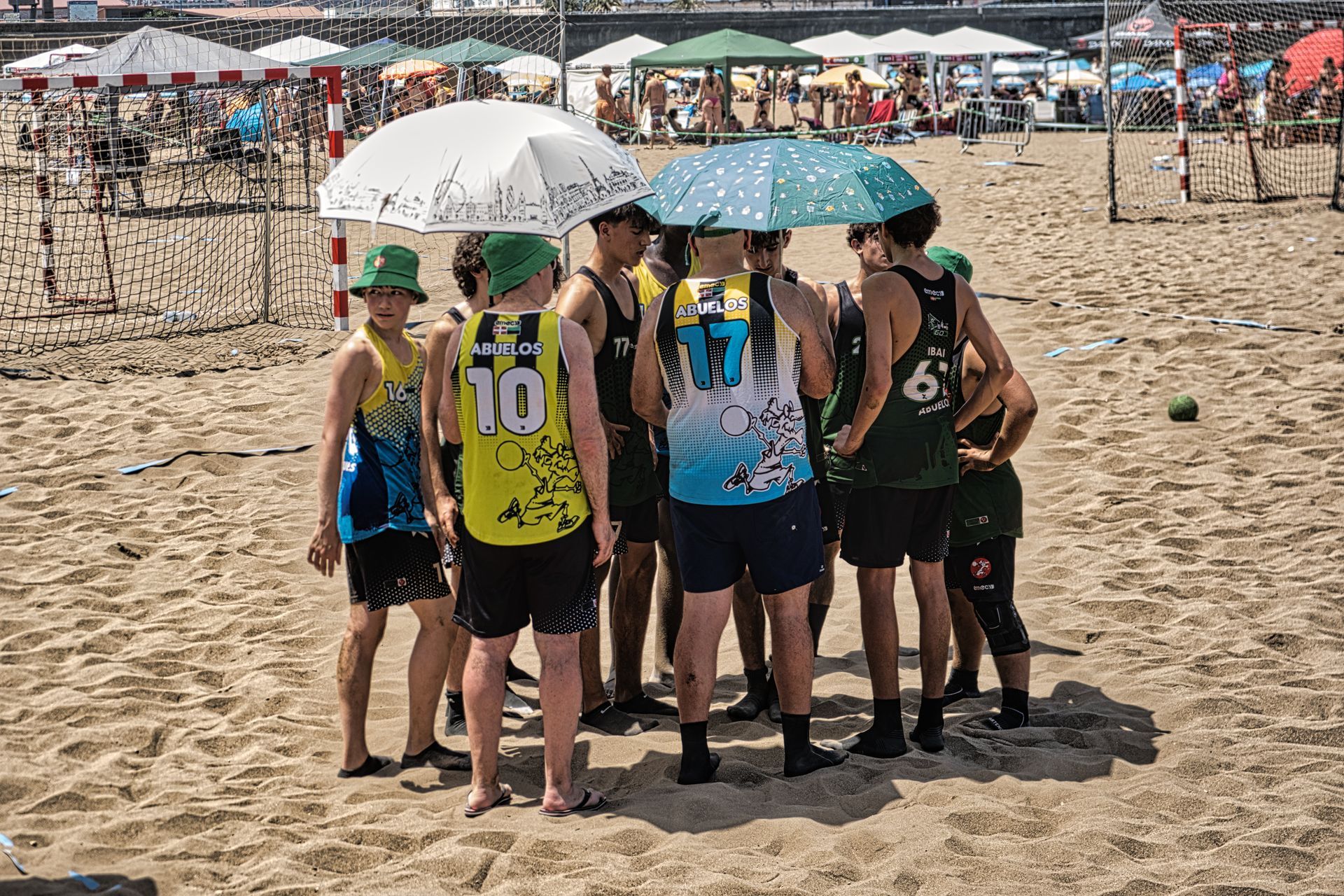 Group of beach athletes huddled under umbrellas on a sandy field, wearing numbered jerseys.