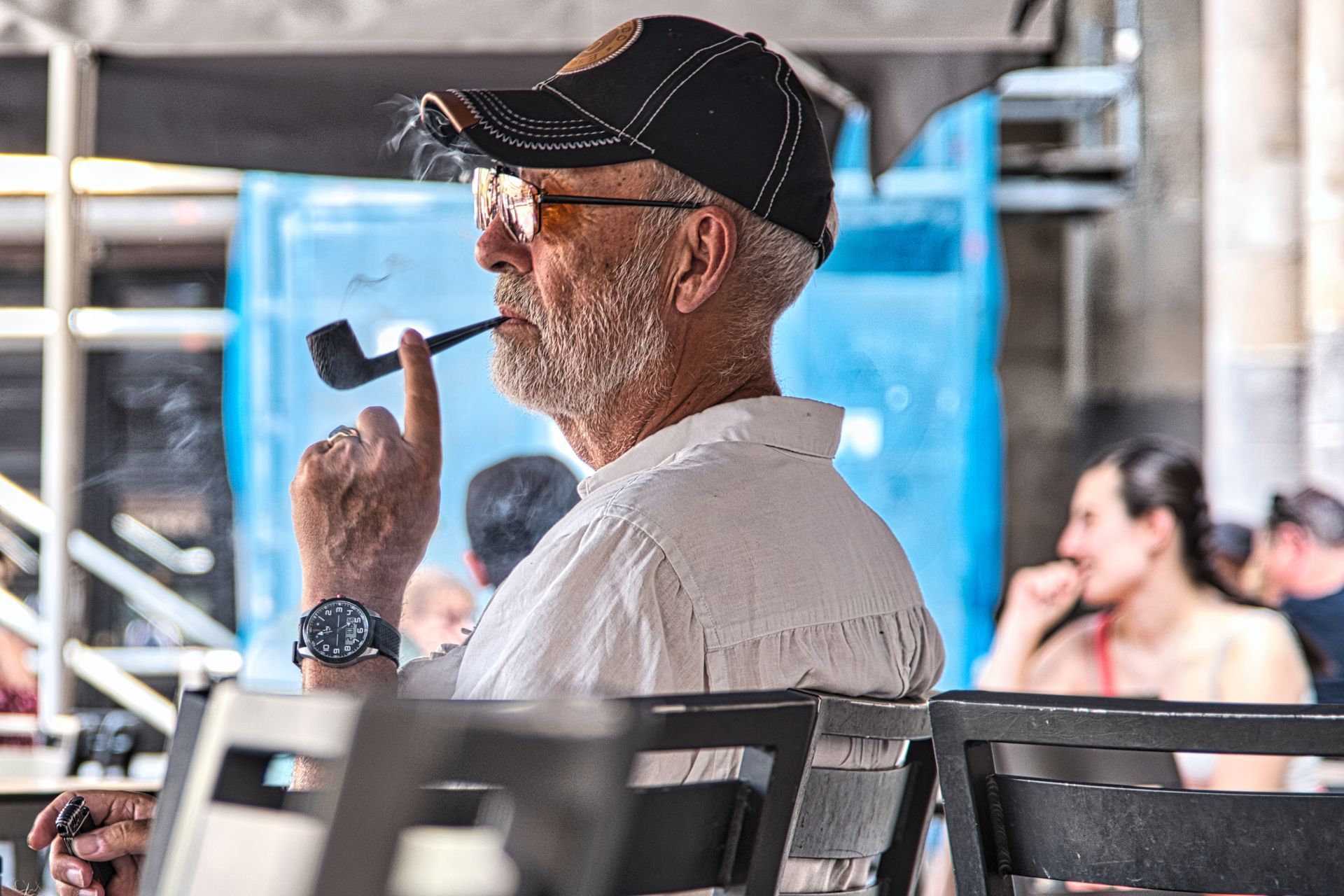Man in a cap smoking a pipe while seated in an outdoor café, with blurred people in the background