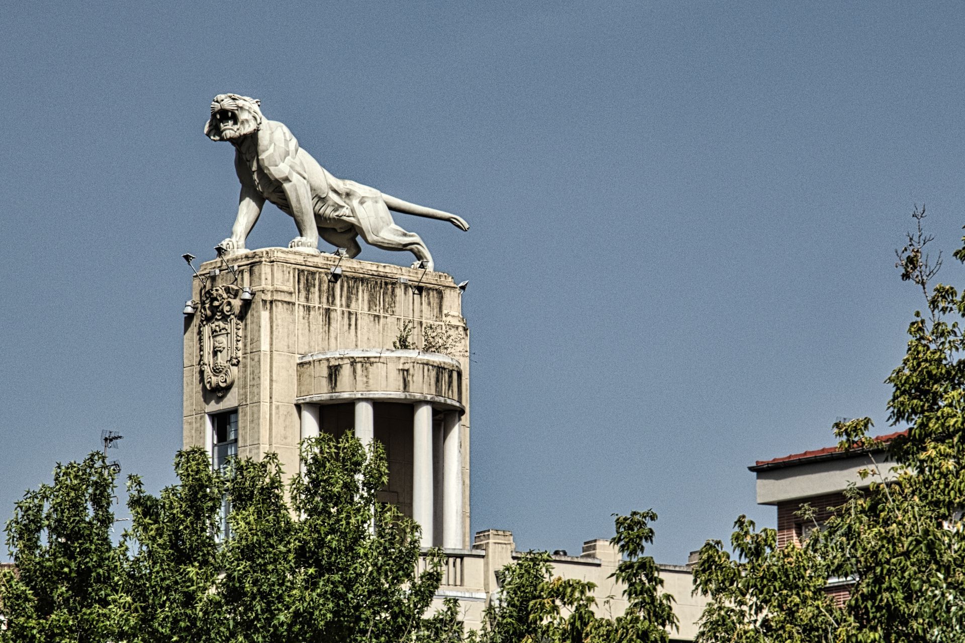 White tiger statue atop a stone tower above green trees under a clear blue sky