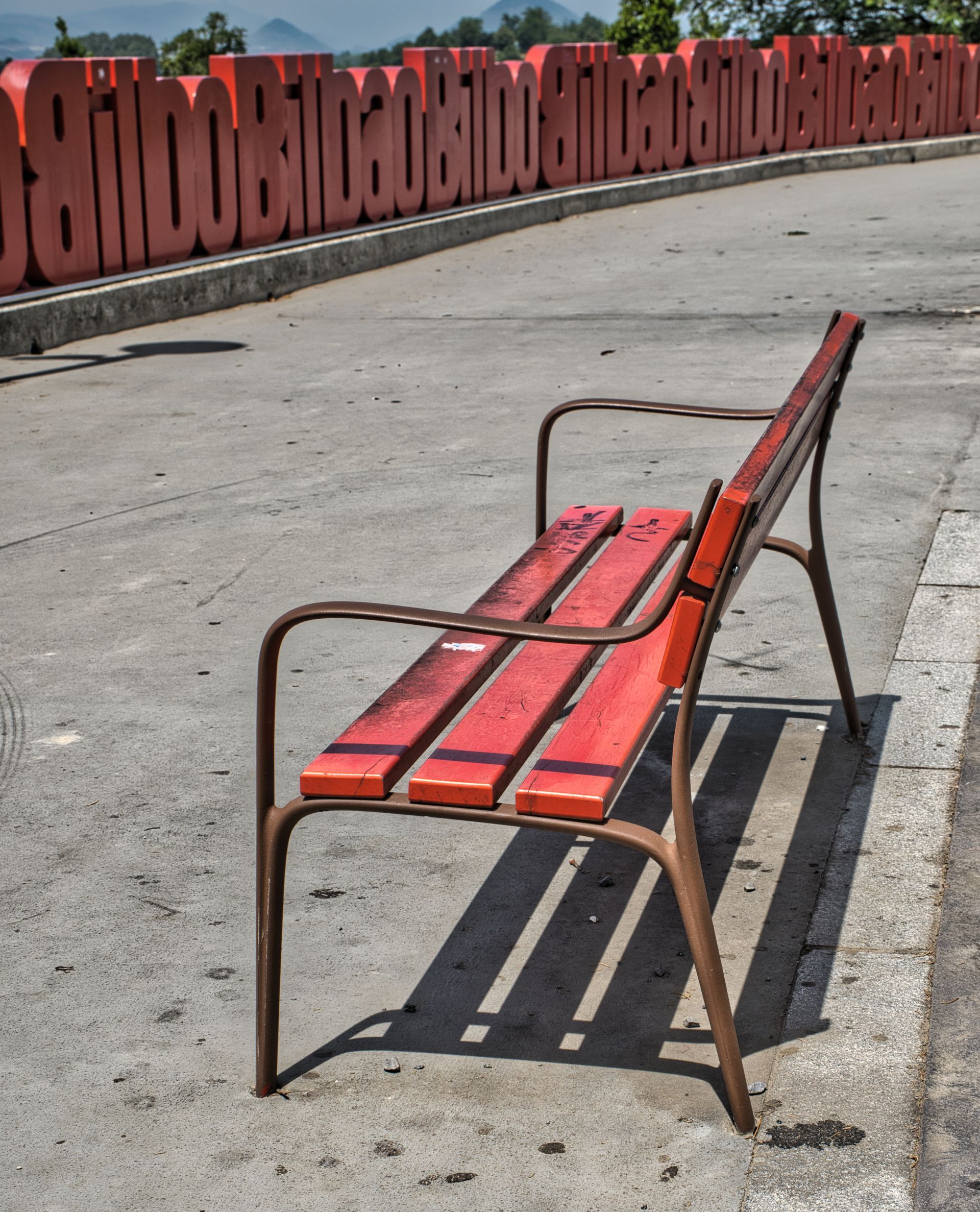 Red park bench on a sunny concrete walkway, with long shadows and a red fence in the background.
