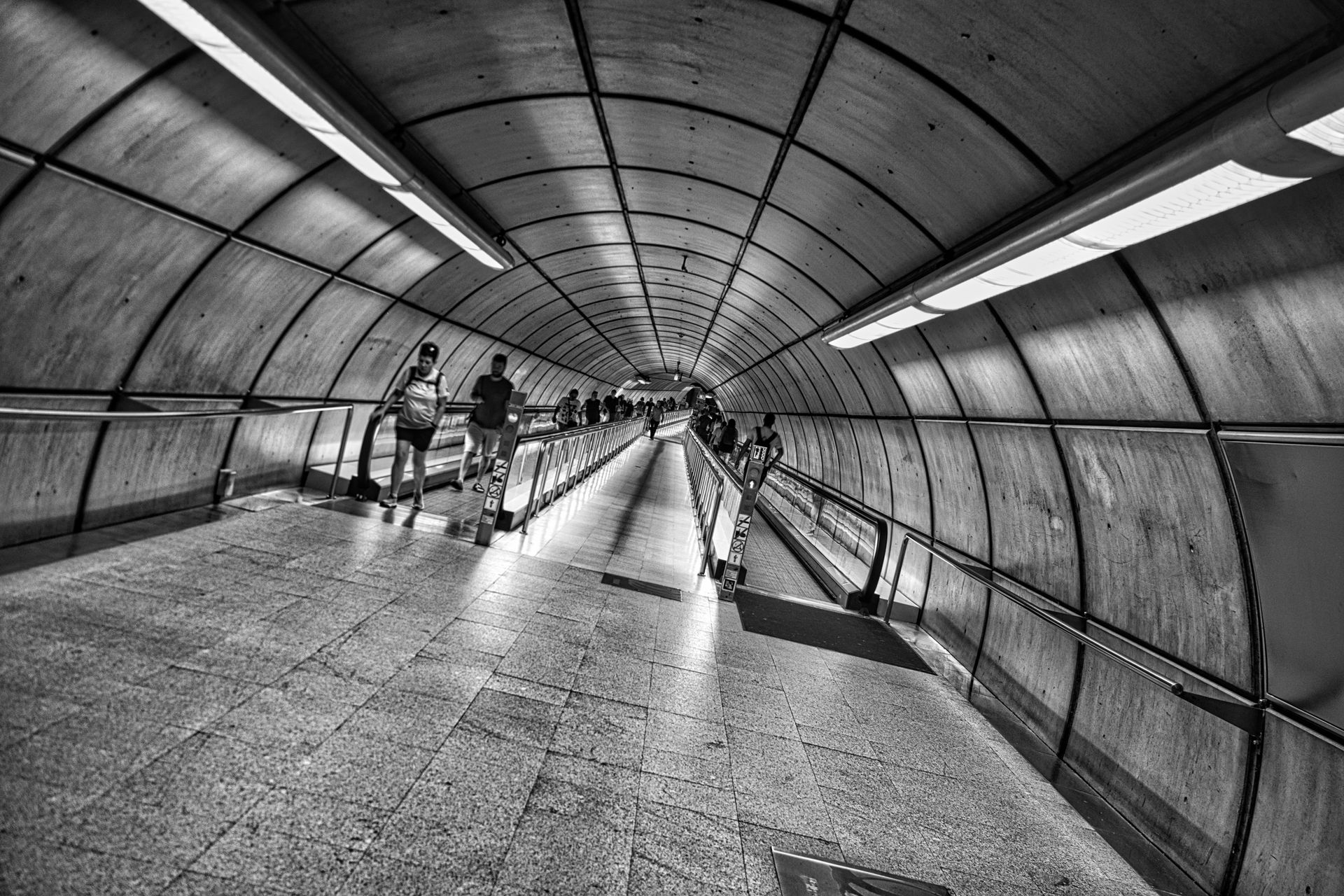 Black-and-white view down a tiled pedestrian tunnel with several people walking through it