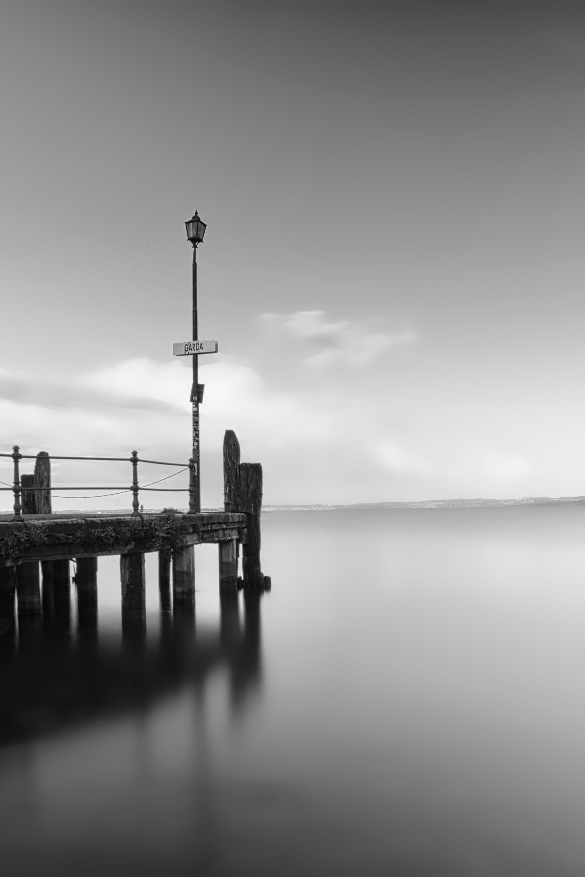 Black-and-white view of a calm harbor pier with a tall light pole and reflections on still water