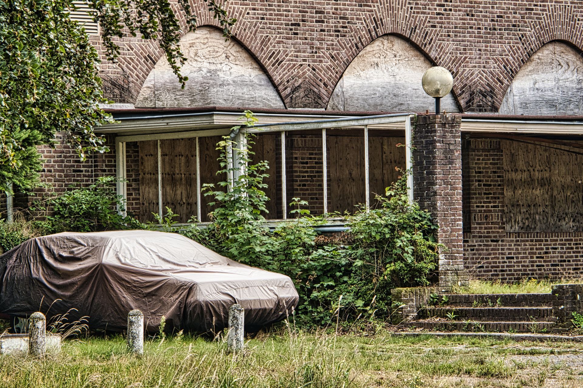 Car covered with a tarp in front of an old brick house with ivy and a small porch